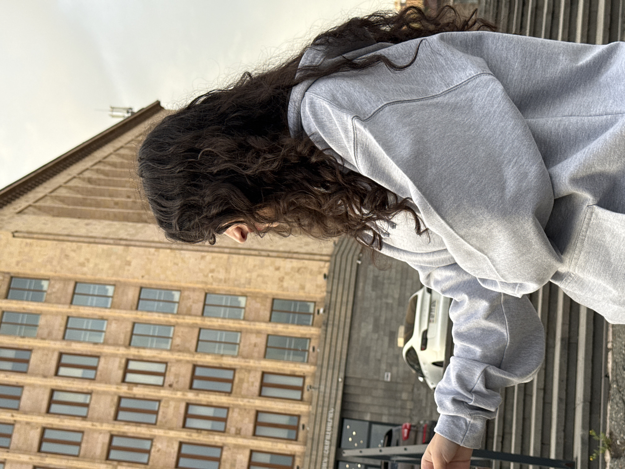A low-angle, medium shot depicts a person with long, dark curly hair wearing a light gray hooded sweatshirt. The individual is facing downwards and slightly to the right of the frame. In the background, a multi-story building with numerous symmetrically arranged windows is visible. To the right of the building, there is a street scene with a white vehicle, a concrete wall, and other urban infrastructure. The photograph was taken in Yerevan, Armenia.