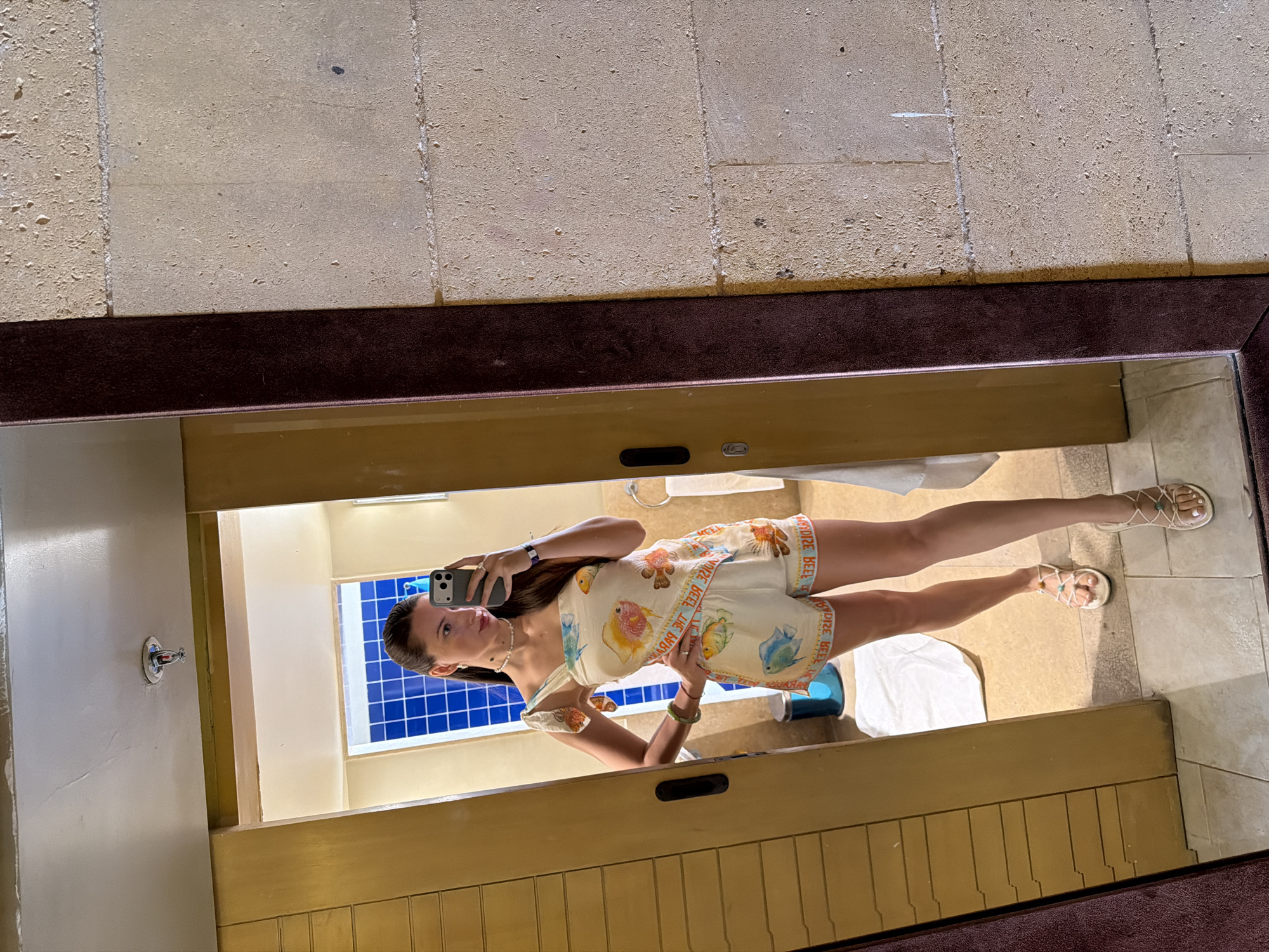 A full-body, front-facing mirror selfie shows a woman standing in a bathroom. She is wearing a white, sleeveless, one-shoulder top and matching shorts, both adorned with a pattern of colorful fish and text. Her hair is pulled back in a ponytail, and she is wearing a pearl necklace and earrings. The bathroom features tiled walls, a wooden vanity, and a shower with blue tiles. The overall environment suggests a hotel or resort. The photographer is holding a black smartphone in her right hand.