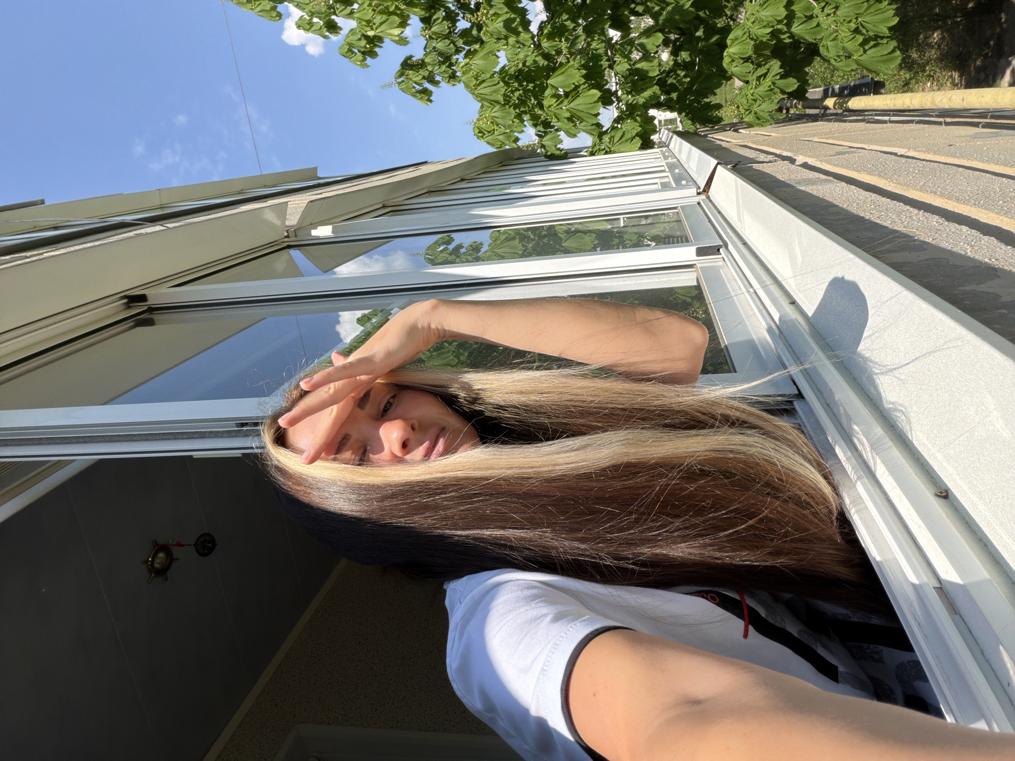 A woman with long brown and blonde hair is leaning out of a window. She is wearing a white t-shirt. She has her hand raised to shield her eyes from the sun. The window is located in a multi-story building. Outside, there is a tree with green leaves and a section of a concrete roof with a yellow pipe. The location is the city of Kamyshin, Russia.