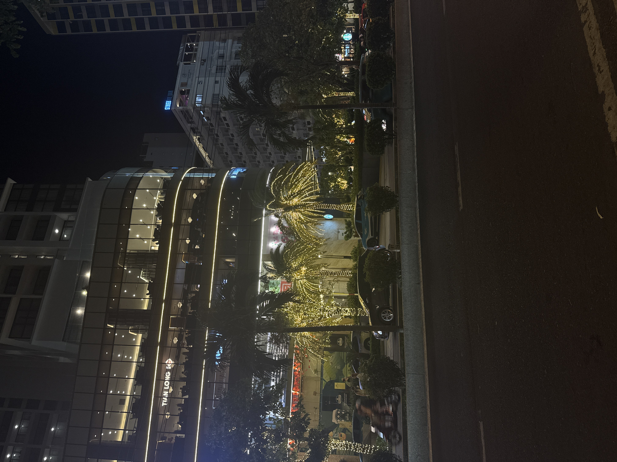 At night, the city of Nha Trang Ward, Vietnam is illuminated with lights. A multistory building with a glass facade displaying the words "TIAN LONG BO" is visible. Palm trees decorated with string lights line the street in front of the building. Cars are parked along the side of the road. A tall building is partially visible in the background. A road occupies the right side of the frame.