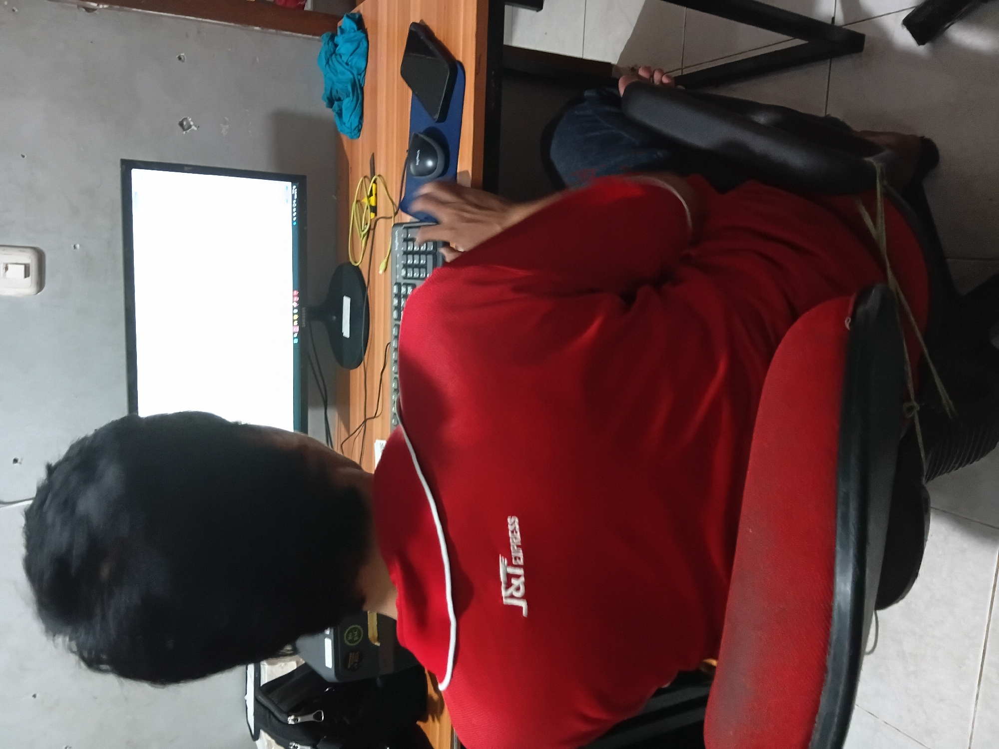 A person sits in a red office chair, facing away from the camera, and working on a desktop computer. The person is wearing a red polo shirt with "J&T EXPRESS" embroidered on the back in white lettering. The computer monitor is on, displaying a white screen, and a keyboard and mouse are on the desk in front of it. To the right of the mouse, a smartphone is lying on a blue mousepad. A crumpled teal cloth is also visible on the desk. The desk is made of light brown wood, and the wall behind it is gray and has visible marks and holes. To the left of the monitor, a black bag is placed on top of a printer. The scene appears to be an indoor workspace, possibly an office or a home office. The flooring is tiled. The image was taken in Bekasi, Indonesia.