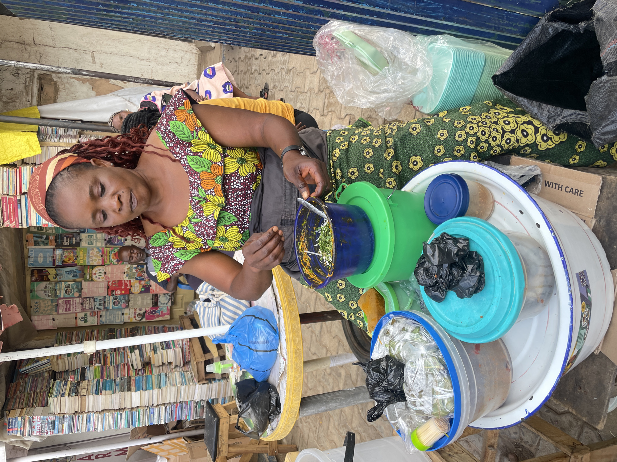 In Jos, Nigeria, a woman with red-tinged hair and a patterned headband wears a floral-printed top and a dark gray apron. She is preparing food, possibly 'awara' a popular local snack, using a blue bowl and a spoon. Several plastic containers of various colors and sizes surround her, sitting on top of a large metal bowl. A collection of books is visible in the background, suggesting she might be working near a bookstore. Another person is seated behind the books. Additional people are lying on mats nearby, possibly resting. A yellow table and various plastic bags and containers are scattered around the area.