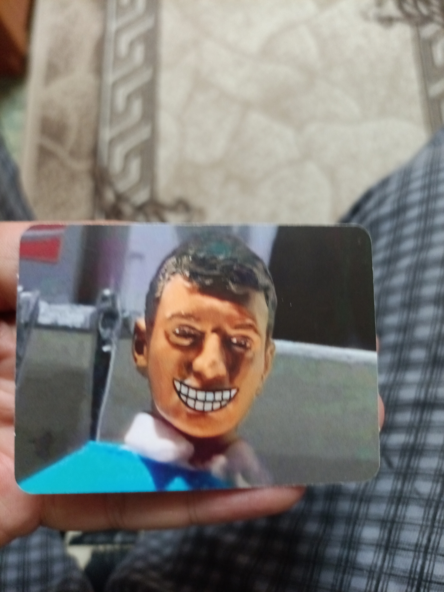 A close-up shot shows a person holding a card with an image of a doll's head on it. The doll has dark hair, a tan complexion, and a wide, toothy grin. The background of the card is blurred, but appears to show some metallic structures. The person holding the card is wearing a plaid garment. The location of this image in Nyagan, Russia is unknown.