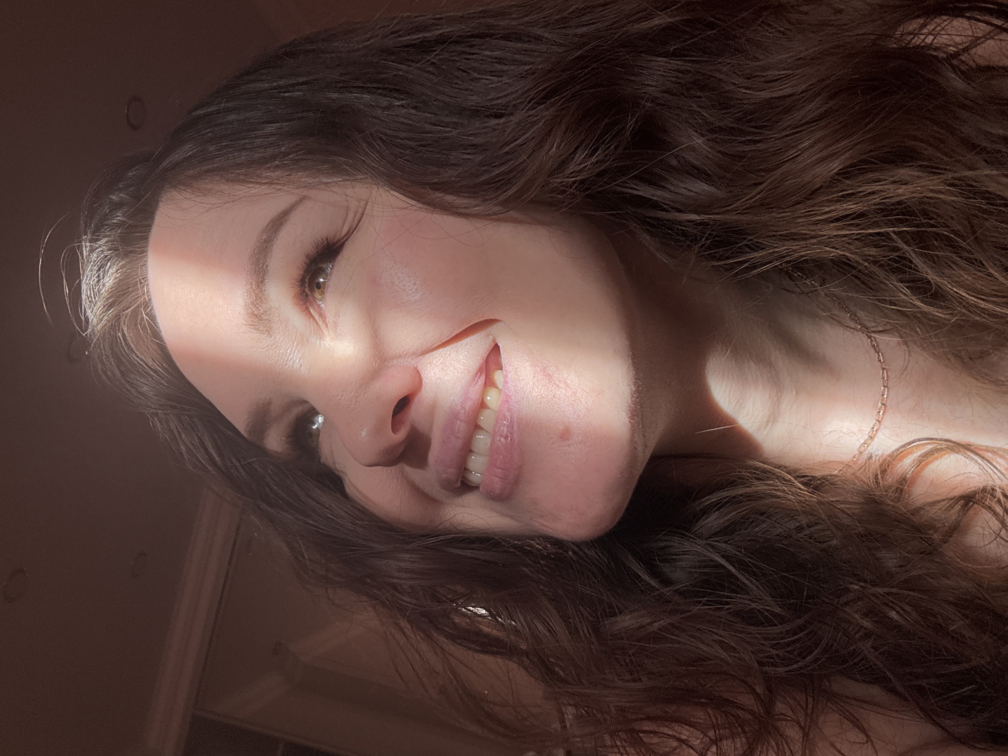 A young woman with long, wavy dark brown hair is captured in a close-up, angled portrait, likely in Yuzhny, Russia. The image is rotated, presenting her face on a diagonal tilt, suggesting she might be lying down or leaning back. A strong, warm diagonal beam of sunlight illuminates the left side of her face, highlighting her light brown or hazel eye, nose, and a portion of her smiling mouth, which reveals healthy teeth. Her eyebrows are well-defined, and she has long eyelashes.
Her expression conveys a sense of gentle happiness or contentment. Her dark hair cascades around her, partially obscuring her neck and shoulders, where a delicate gold-colored chain necklace is visible. The rest of the scene is cast in shadow, with only faint outlines of what appears to be an indoor setting, possibly a wall or furniture, in the background. The distinct sunlight suggests either an early morning or late afternoon time of day. There is no visible text in the image.