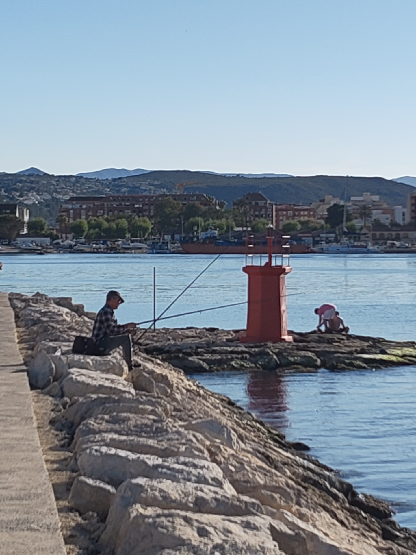 This daytime photograph captures a serene coastal scene in Dénia, Spain, under a clear blue sky. The main subjects are a man fishing and two individuals near a small red lighthouse, set against a backdrop of a bustling harbor, a town, and distant mountains.
In the foreground, a light-colored concrete walkway runs along the left edge, leading to a rugged breakwater made of large, grey rocks. A man, wearing a plaid shirt and a cap, is seated on these rocks, actively engaged in fishing with two rods positioned in the water. Behind him, a black bag or tackle box rests on the rocks.
To the right, a small red cylindrical lighthouse, acting as a navigational aid, stands prominently on a cluster of dark, mossy rocks in the water. Next to it, two people are seen bent over, seemingly exploring the waterline or the rocks. One person appears to be an adult in a light-colored shirt, possibly assisting a child. The calm, blue water around the lighthouse reflects its red color faintly.
The midground features a body of water, likely a harbor or marina, dotted with several boats. In the background, a coastal town spreads across the land, characterized by various buildings and green trees. Further back, undulating mountains complete the horizon, fading slightly into the distance.
The overall impression is one of a peaceful, sunny day by the sea, with people enjoying leisure activities in a scenic Mediterranean setting. There is no visible text in the image.