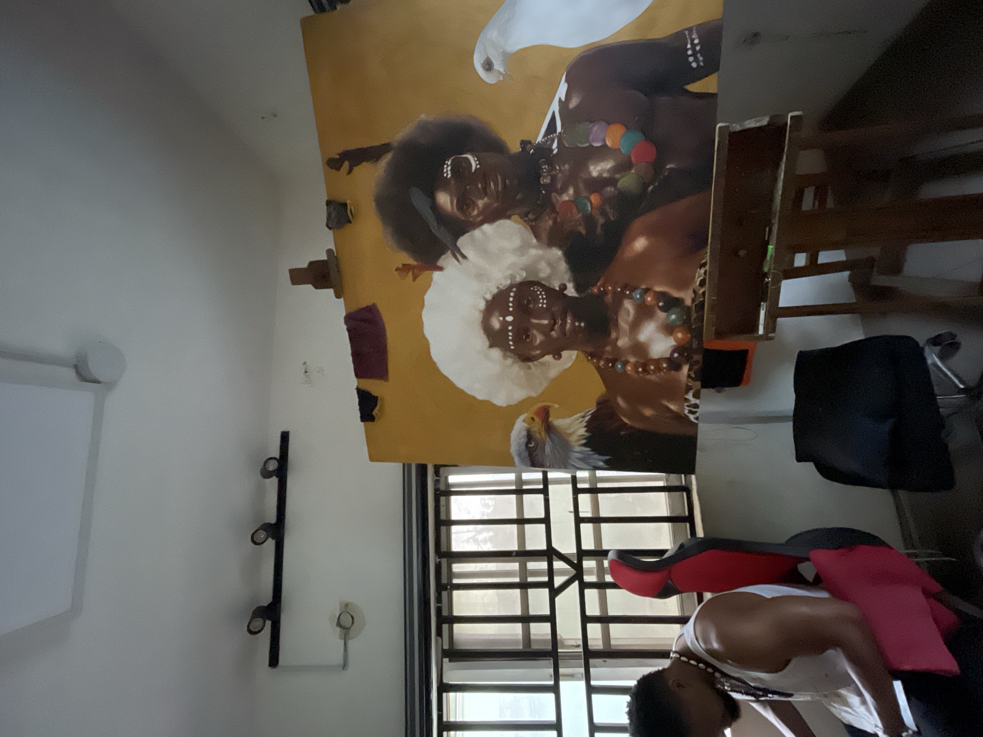 In a studio space in Ado, Nigeria, a man wearing a white tank top and necklaces is seated on a red and black gaming chair. He is looking down at a tablet or book in his lap. To his right, a large painting is displayed on an easel. The painting depicts an eagle, a man with an afro and tribal markings, a woman with a large white afro and tribal markings, and a white dove. The studio has a window with barred security and is lit by track lighting on the ceiling. A wooden desk with art supplies sits in the foreground.