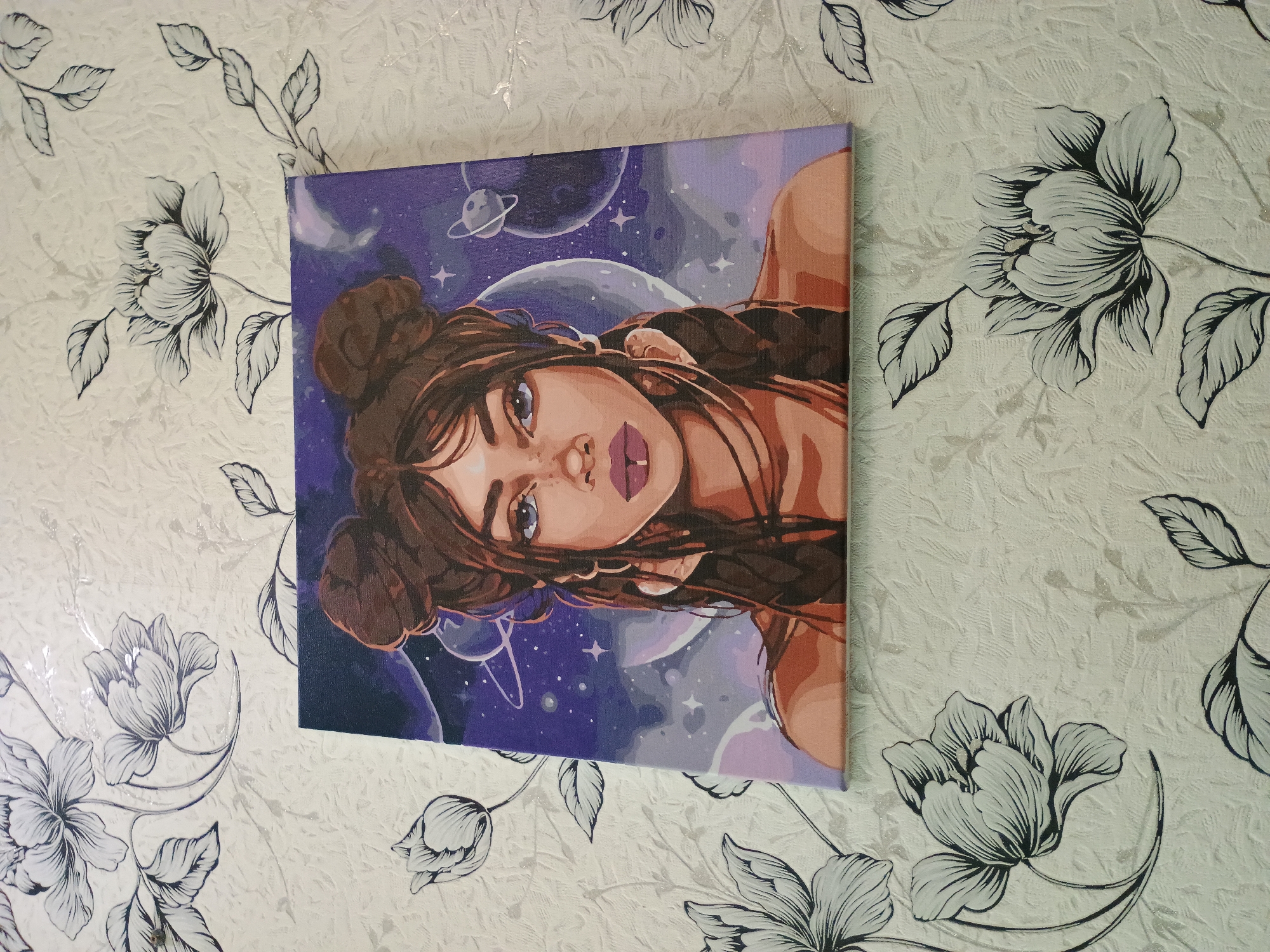 A square painting depicting a woman with brown hair styled in two buns and two braids against a purple, star-filled sky with planets is laid on a white tablecloth with a black floral pattern. The painting is positioned slightly to the left of the center of the frame, with its bottom edge closer to the viewer. The woman has light brown skin, full lips with purple lipstick, and large, dark blue eyes. She wears large hoop earrings and a septum ring. The background of the painting features several planets and celestial bodies. The tablecloth has a textured surface with a repeating pattern of large, stylized flowers and leaves. The lighting appears to be natural, casting soft shadows. The overall scene is a still life photograph of the painting on the tablecloth.