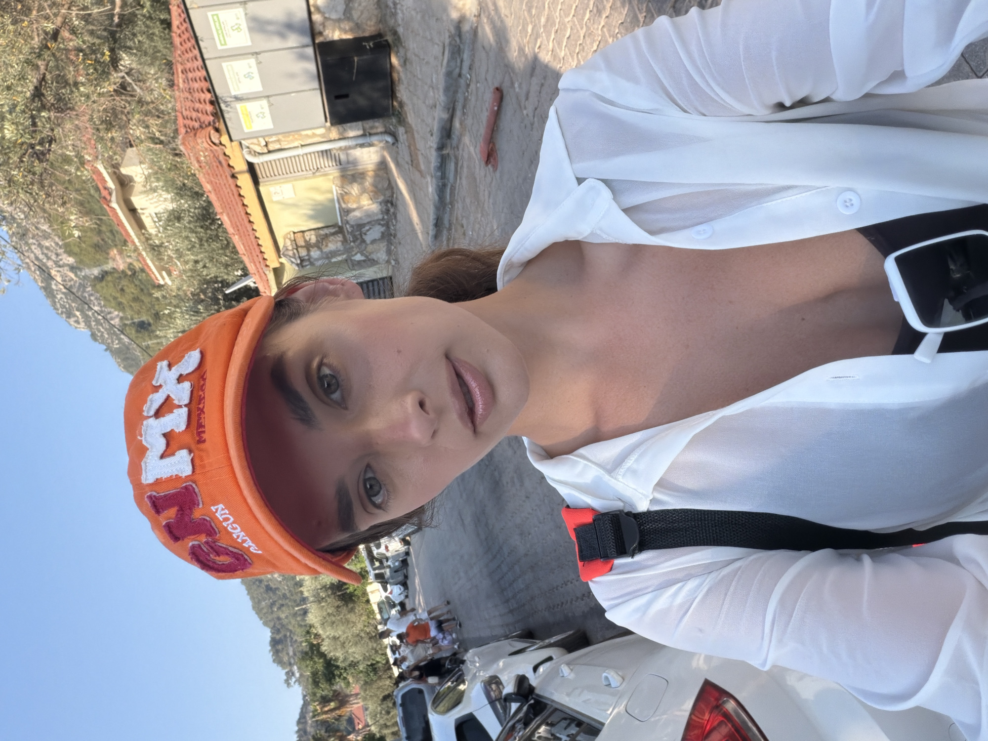 A woman wearing an orange baseball cap and a sheer white button-up shirt is taking a selfie outdoors. The cap has "GN MX" embroidered in red and white lettering. She is also wearing a dark-colored top underneath her shirt and sunglasses are visible hanging from her shirt. In the background, a street is lined with parked cars and a few people are walking. Buildings with red tile roofs and trees are visible behind the street, and a mountainous landscape can be seen in the distance. The scene appears to be in a sunny, urban environment in Fethiye, Turkey, likely on a street with shops or residences.