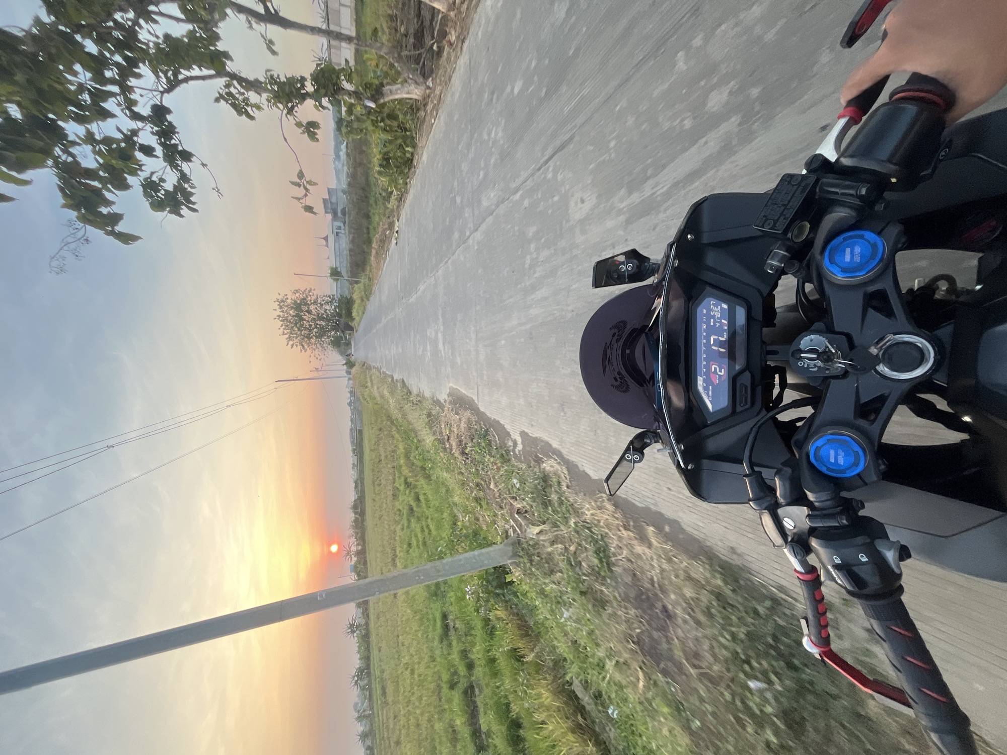 The image is a first-person perspective of someone riding a motorcycle on a paved road in Pecangaan, Indonesia. The sun is setting, casting a warm orange glow across the sky and illuminating a field of green crops to the left of the road. A utility pole stands prominently on the left side of the road, with power lines stretching across the sky. Trees and some buildings are visible in the distance. The motorcycle's handlebars, dashboard displaying speed and other information, and the rider's hands gripping the handlebars are in the foreground. The road appears to be relatively empty, with no other vehicles in immediate view. The overall impression is one of a peaceful sunset ride through a rural area.