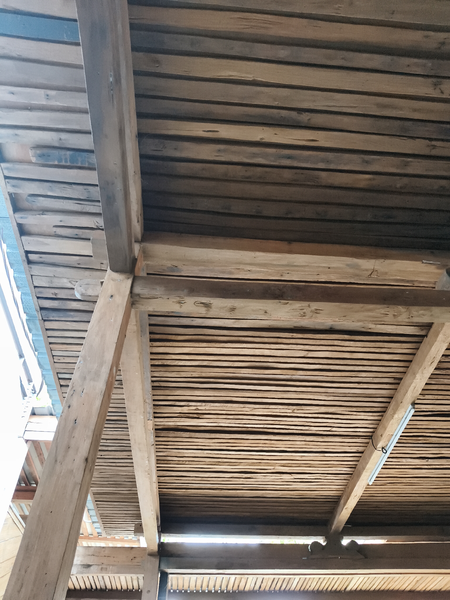 The image shows the underside of a wooden roof structure, likely part of a building in Nologaten, Indonesia. The roof is constructed from numerous thin wooden planks laid parallel to each other, supported by thicker wooden beams and posts. The wood appears weathered and aged, with variations in color and texture. Some planks show cracks and irregularities. The structure is viewed from below, with beams running diagonally and horizontally, creating a sense of depth and construction. Natural light filters through gaps in the planks in some areas, suggesting an outdoor or semi-open space. There are no people or animals visible in the image. The overall impression is of a rustic and functional wooden architecture.