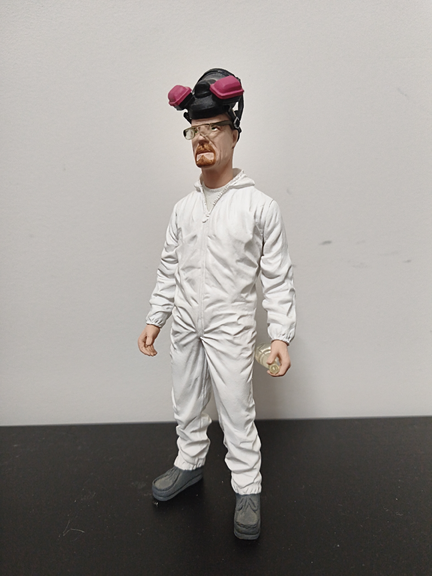 The media file depicts a full-body, front-facing shot of a Walter White action figure, a character from the television series *Breaking Bad*. The figure is standing on a dark, flat surface against a plain white background. It is wearing a white hazmat suit, complete with a zipper down the front, elastic cuffs, and gathered ankles. On its head, the figure sports a black gas mask with two pink respirator cartridges. The figure's face is detailed with glasses, a red goatee, and an open mouth. In its right hand, the figure is holding a small, clear glass vial. The lighting suggests a controlled indoor environment, and there are no visible environmental cues to indicate the time of day or weather. The setting appears to be a studio or display area, with no discernable location context beyond what is suggested by the *Breaking Bad* character. No text is visible in the image.