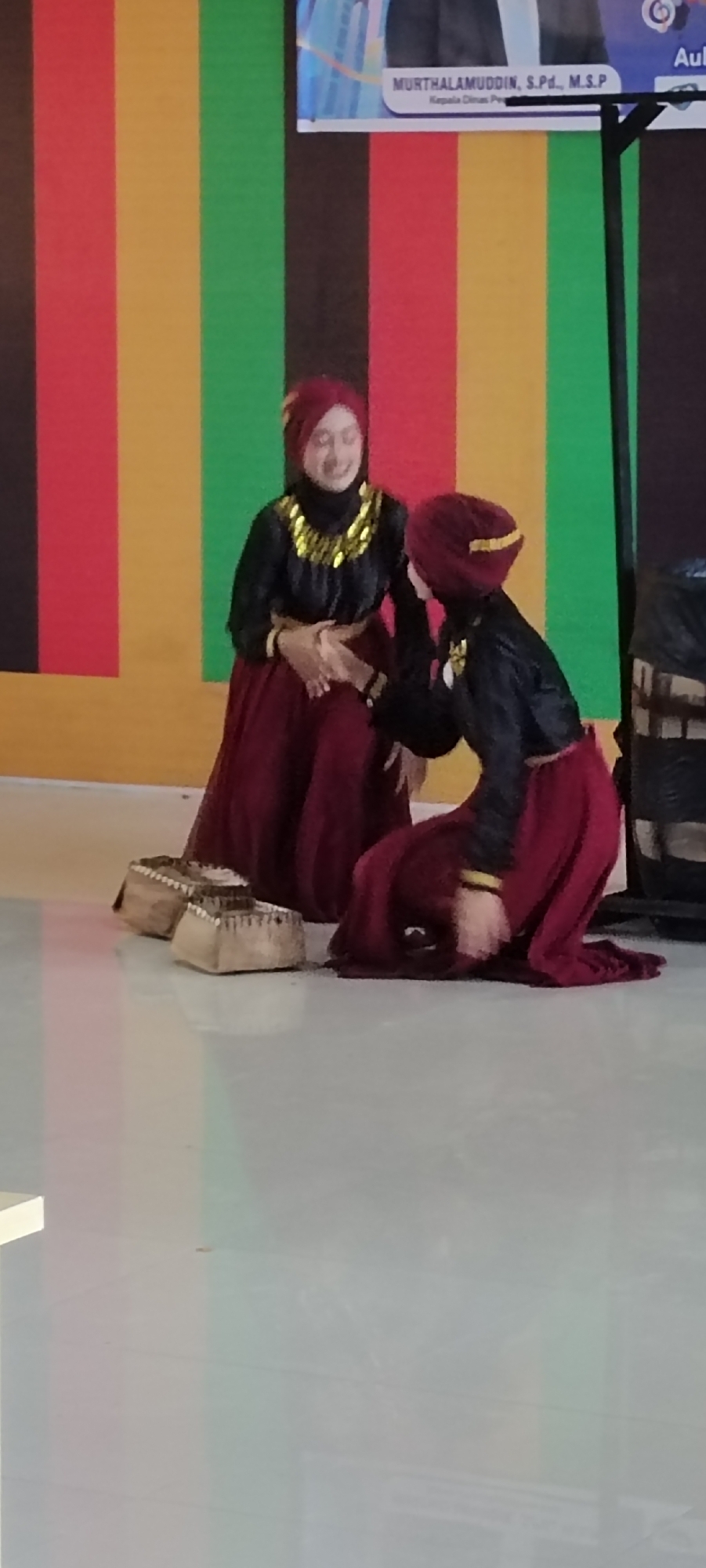 Two women are performing a traditional dance on a polished floor, with wooden shoes (likely used as musical instruments) on the left. The woman on the left is sitting, smiling, and holding the other woman's hand. The woman on the right is kneeling and bowing her head. They are both wearing matching outfits consisting of a burgundy skirt and a black blouse with gold trim. A backdrop behind them features vertical stripes of black, red, yellow, and green, and a sign at the top with the name MURTHALAMUDDIN, S.Pd., M.S.P. Kepala Dinas Per. The image was taken indoors, the precise location in the city of Unknown, Unknown, is not discernible.