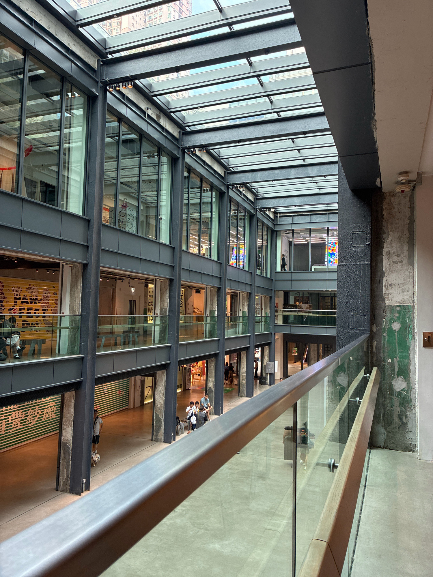 The image depicts the interior atrium of a multi-story building in Hong Kong, China, likely a commercial or mixed-use development. The architecture features a modern industrial aesthetic with exposed steel beams, concrete pillars, and extensive glass paneling, including a large glass roof that allows natural light to flood the space. Several levels of walkways and shops are visible, connected by open spaces and balconies.
On the ground floor, a person is walking a small dog. Further in the distance, a group of people are sitting on a bench. On upper levels, various shop fronts with glass windows are visible, some displaying merchandise or signage. One shop has a distinctive yellow facade with red lettering. Another upper floor window shows a person walking past a colorful art installation.
The railings in the foreground are made of metal and glass, with a wooden accent on the lower section of the handrail. The overall environment suggests a public indoor space, possibly a shopping mall or a creative hub, with a clean, contemporary design. The lighting is a mix of natural light from the atrium roof and artificial lighting within the shops. The condition of the concrete walls suggests a deliberate raw, unfinished look as part of the design.