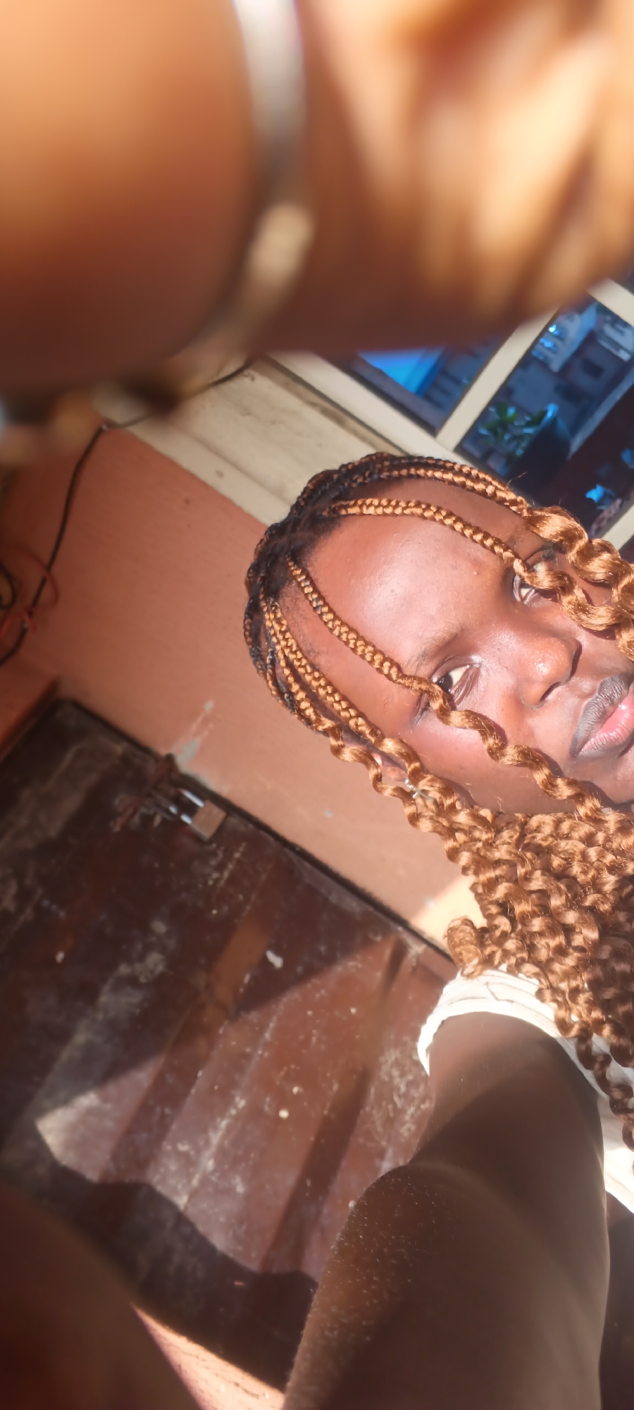 A tilted selfie shows a young woman with dark skin and braided hair extensions, styled in cornrows at the scalp and falling in reddish-brown curls around her face. A warm, golden light illuminates the left side of her face and hair, suggesting late afternoon or early evening. She is wearing a white top, visible on her right shoulder, and looks directly at the camera with a neutral expression.
The top left of the frame is obscured by a blurry hand or arm, likely holding the camera, on which a silver ring is visible. The background features a reddish-brown wall. To the upper right, a window with a white frame shows a blue light source, possibly artificial light from an adjacent room or the ambient light of twilight, along with hints of distant structures. Below the window, a dark brown surface, possibly a wooden door or furniture, appears dusty, and a small padlock is visible. A black cable runs vertically on the wall in the upper left. The scene is set indoors, possibly in Onitsha, Nigeria.