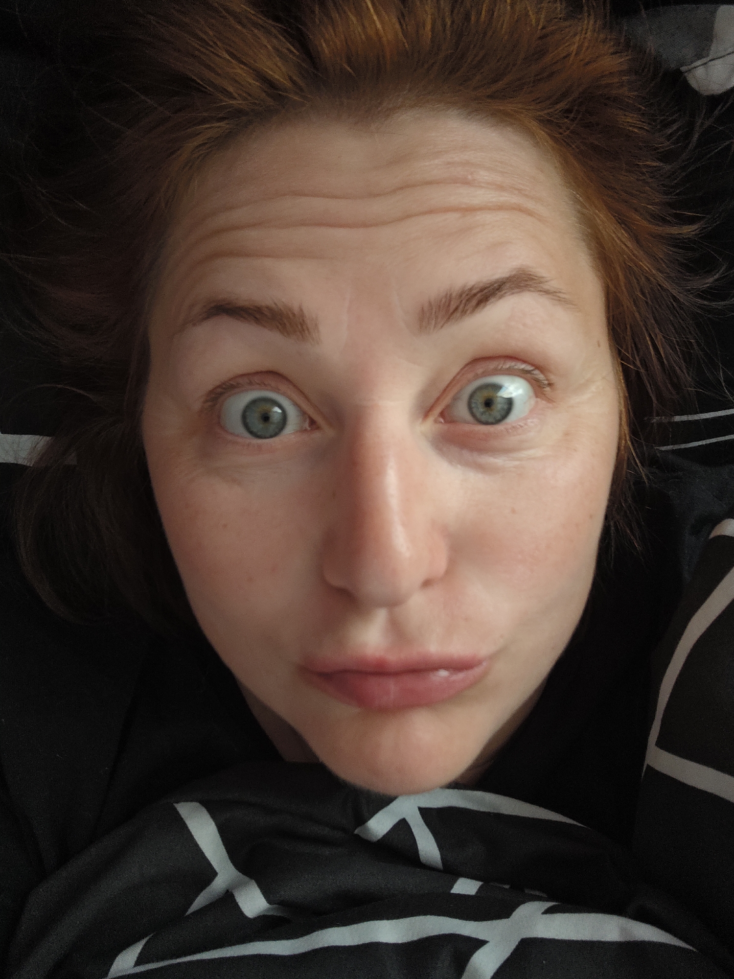 A close-up selfie of a woman with red hair. She has light skin, green eyes, and pursed lips. Wrinkles are visible on her forehead and around her eyes. She is lying on a black and white patterned fabric. The image does not provide enough information to determine her location within Markovskii, Russia, or any specific details about the environment beyond the immediate surroundings.