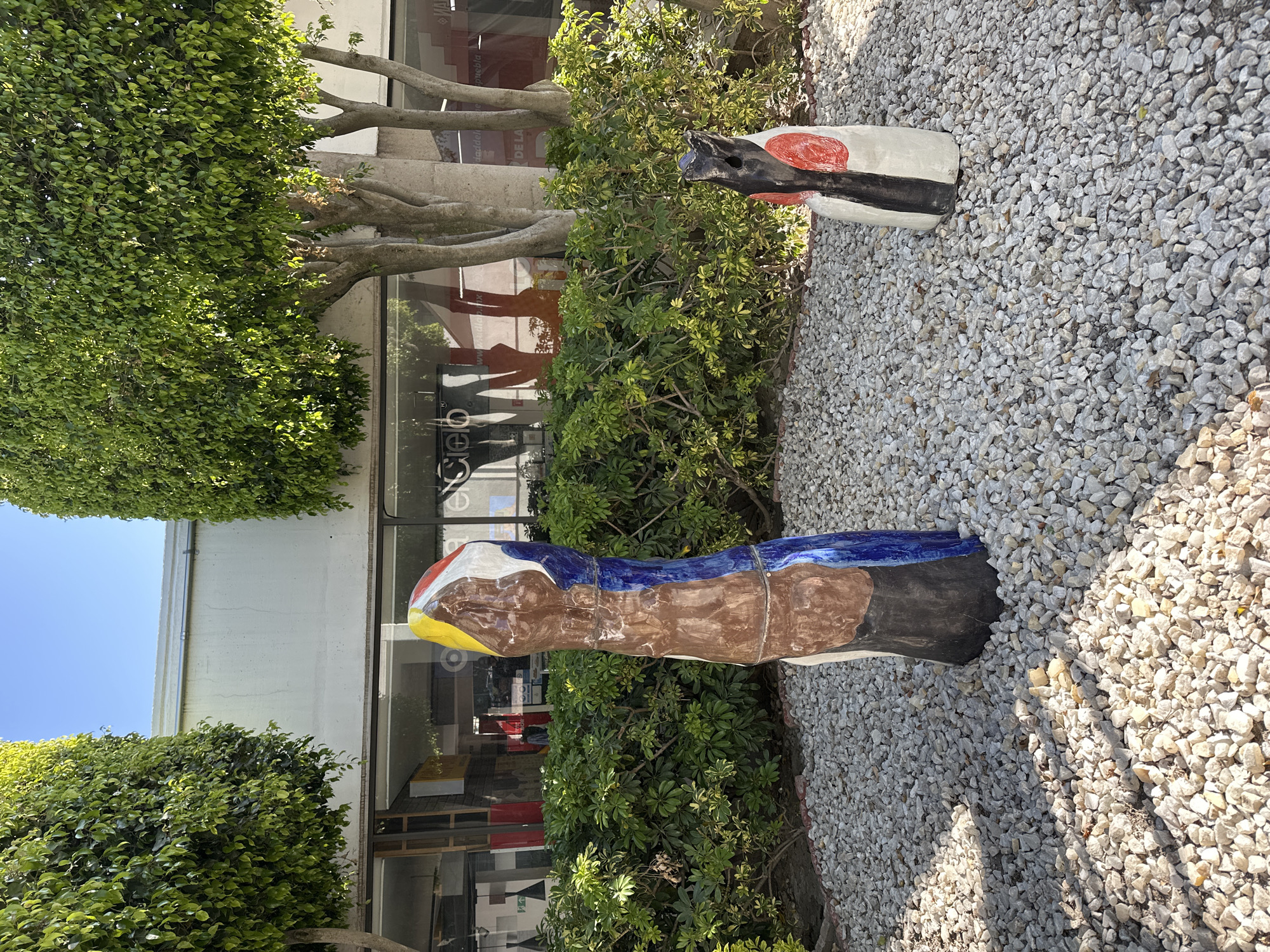 The scene depicts an outdoor landscaped area adjacent to a commercial building in San Andrés Cholula, Mexico. The foreground is covered with light grey and white crushed gravel. Two abstract, multi-colored sculptures are positioned on this gravel.
The taller sculpture on the left, approximately 1.5 meters in height, is segmented and features irregular painted sections of yellow, red, white, brown, dark blue, and black. The shorter sculpture on the right, approximately 0.8 meters tall, is predominantly white with a vertical black stripe, a prominent red circular motif on its side, and an irregular dark-colored top.
Behind the sculptures and gravel, a dense, manicured hedge of green bushes runs horizontally. Two mature trees with thick green canopies and visible grey trunks flank the scene, rising above the building. The background consists of the ground floor of a modern commercial building, characterized by a light-colored facade above large, reflective glass storefront windows. These windows display reflections of the trees and partially legible text, including "Cielo," "www.idlap.mx," and "Puebla." The sky above is clear and blue. Bright sunlight illuminates the area, casting distinct shadows from the sculptures and trees onto the gravel. No individuals are present.