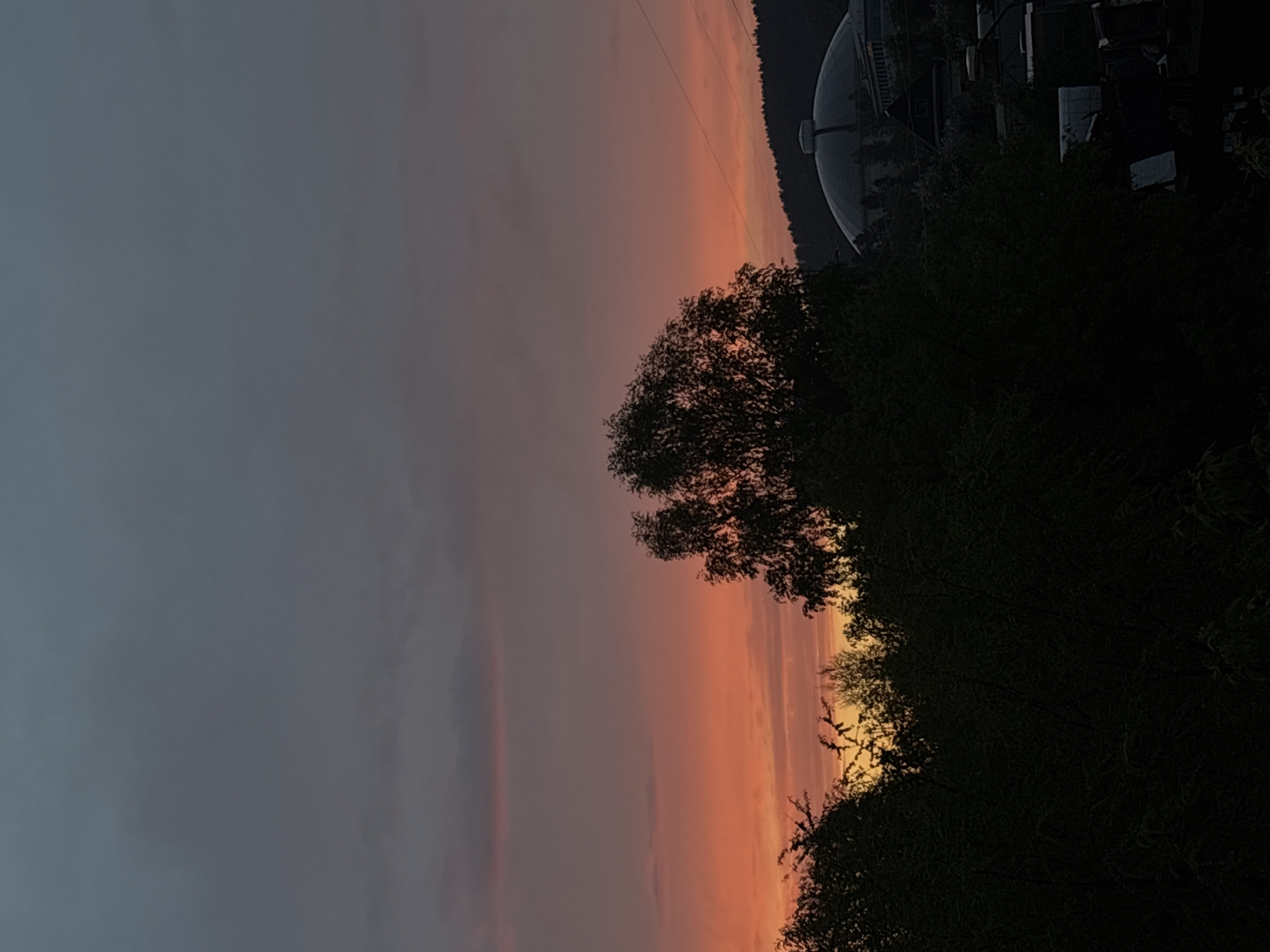 The image depicts a landscape scene, likely at dusk or dawn, in Zheleznogorsk, Russia. A large, spherical structure, possibly a dome or radar, is visible behind trees in the upper right. A dense tree line extends along the right edge of the frame. The sky transitions from dark gray on the left to orange and yellow hues in the center. Overhead power lines are stretched across the upper part of the scene. Silhouette of trees and building are present.