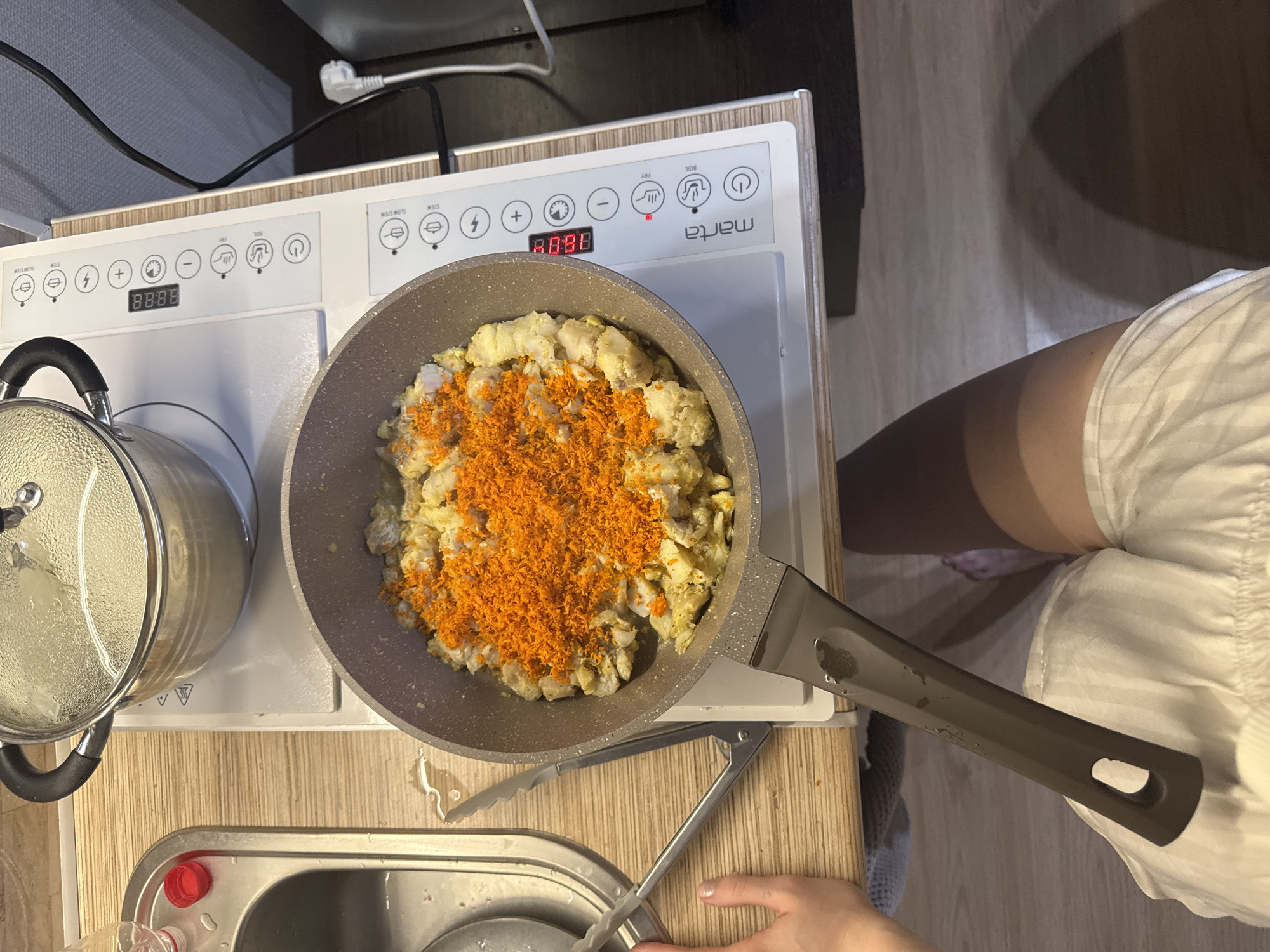 A top-down shot depicts a meal being prepared in a kitchen. A frying pan on a white portable electric cooktop holds diced chicken with shredded carrots. A lidded pot sits beside the pan. Kitchen utensils lie on the countertop near a sink. A person wearing white shorts stands near the cooktop. The cooking area appears to be in an apartment. A "Marta" brand cooktop is visible, suggesting the location could be Russia, specifically the city of Perm, where the brand is available.