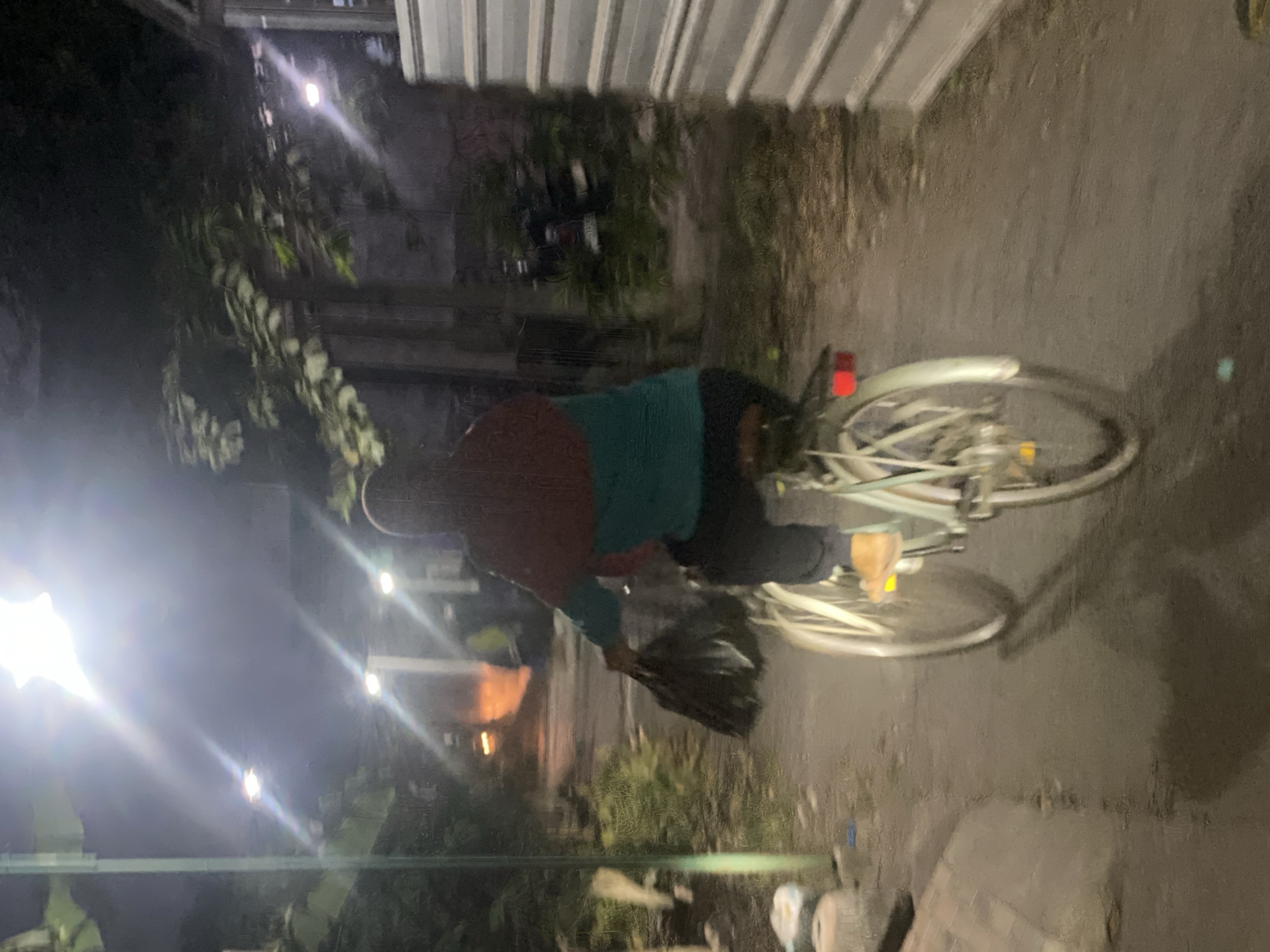 A person is riding a bicycle at night on a wet, paved path in a residential area. The person is wearing a dark hooded garment with a maroon colored fabric draped over their shoulders and a teal colored shirt underneath, and dark pants. They are carrying a large black plastic bag in their left hand, which is hanging down towards the front wheel. The bicycle is light-colored with visible spokes on both wheels and a red reflector on the rear fender. The environment is dimly lit by overhead streetlights, casting bright lens flares. There are trees with green leaves on the left side of the path, and a building with corrugated metal siding on the right. The ground appears wet and reflects the light from the streetlights. In the background, a motorcycle is parked near some bushes. The scene is likely located in the city of Kasihan, Indonesia, given the typical lighting and urban elements present.