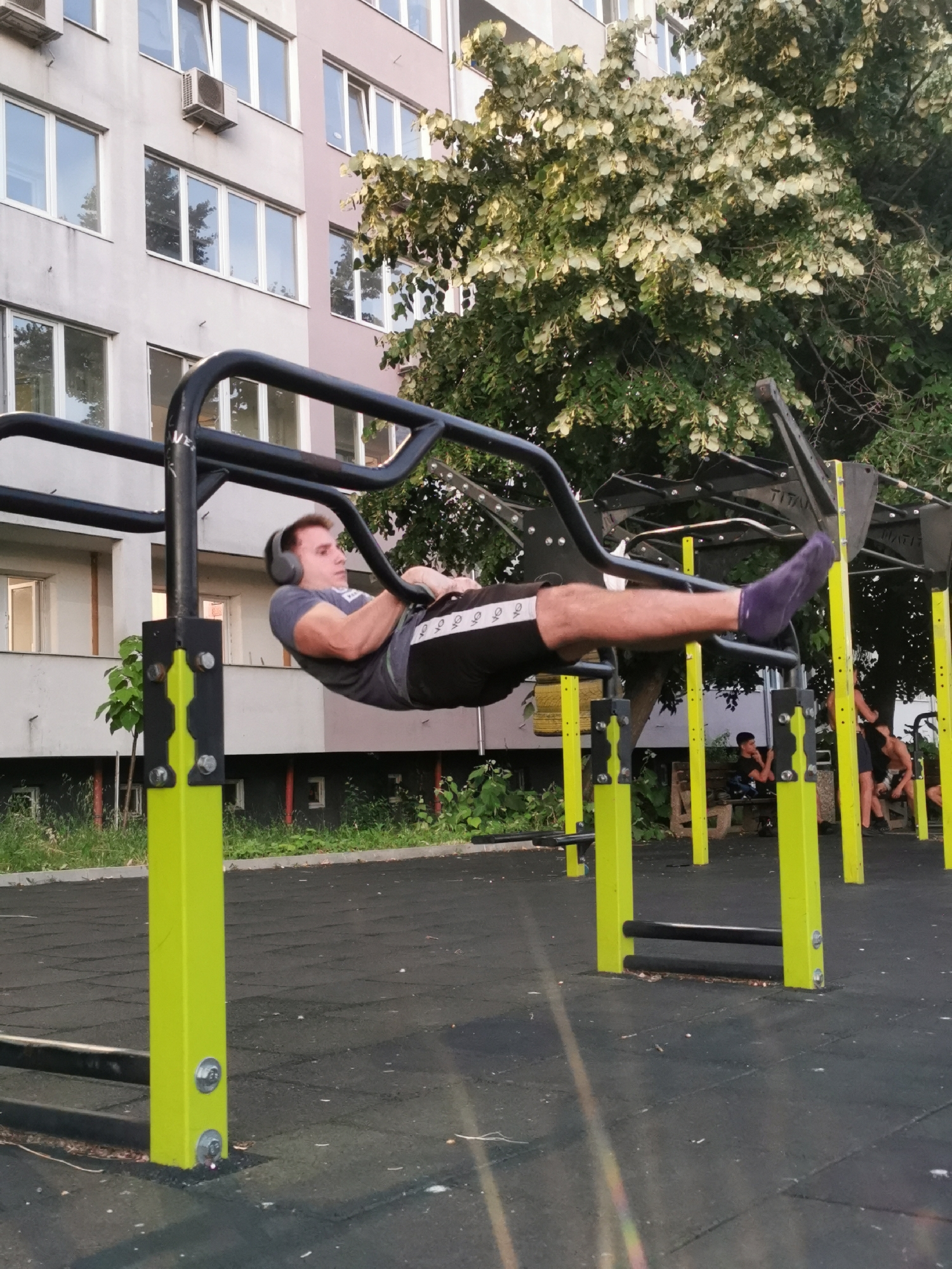 A Caucasian male wearing headphones, a grey t-shirt, and black shorts with white stripes is performing a lever exercise on a black pull-up bar apparatus. His body is horizontal, with his legs extended and feet pointed. The apparatus is bright yellow and black, with a black rubberized ground surface. In the background, a multi-story building with numerous windows is visible, along with a tree with light-colored leaves. To the right, two individuals are seated on a bench, and another person stands. The scene is an outdoor gym in the city of Burgas, Bulgaria.