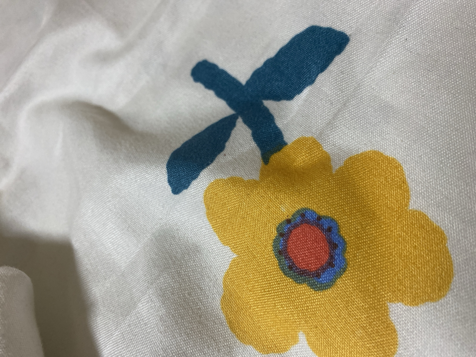 The image is a close-up photograph of a white fabric patterned with a stylized floral design. The fabric itself is soft and shows visible wrinkles and folds, particularly on the left side and top of the frame. The textile exhibits a fine, plain weave texture, which is visible even through the printed areas.
The main subject of the pattern is a vibrant yellow flower with roughly eight rounded petals. Its center features a circular red shape, which is encircled by a scalloped blue ring, further outlined by a thinner dark blue border. Above and to the left of the flower, two elongated, dark teal shapes, suggestive of leaves or a stem, cross over each other. The printed colors have a slightly matte finish, and the fabric's texture is discernible beneath the printed pigments.
There is no discernible activity or event taking place, as the image focuses solely on the static textile pattern. No individuals or objects other than the fabric are visible. The lighting appears soft, highlighting the fabric's contours. There is no visible text. While the image itself does not provide location cues, the context states that it was taken in Sofia, Bulgaria.