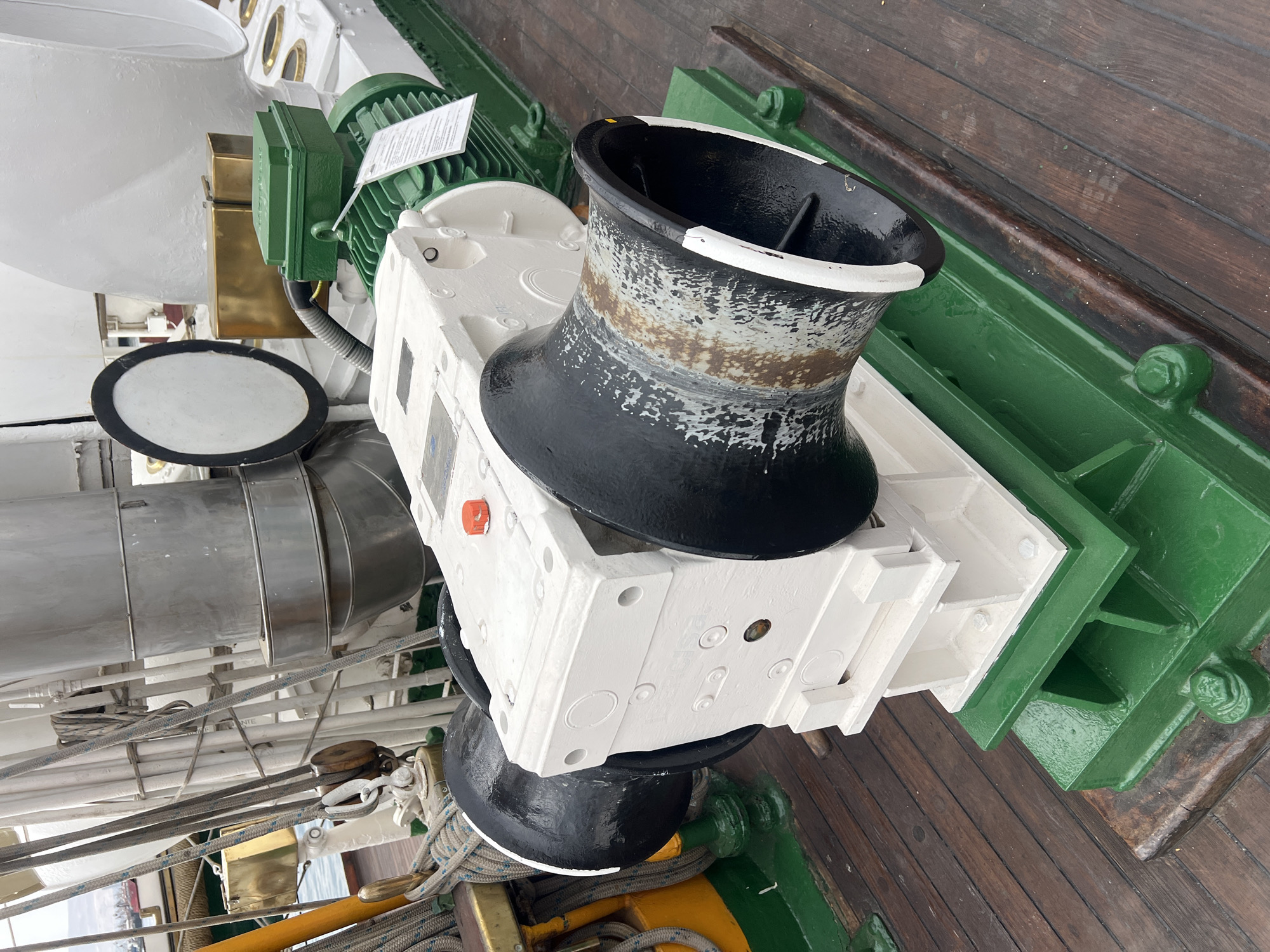 The image shows a close-up of a capstan on a ship, located in Arica, Chile. The capstan is primarily white and black, with some wear and tear visible. It appears to be a part of a sailing ship, sitting on a wooden deck next to green painted metal structures. Other mechanical components like pipes, pulleys, ropes, and other pieces of machinery are in view.