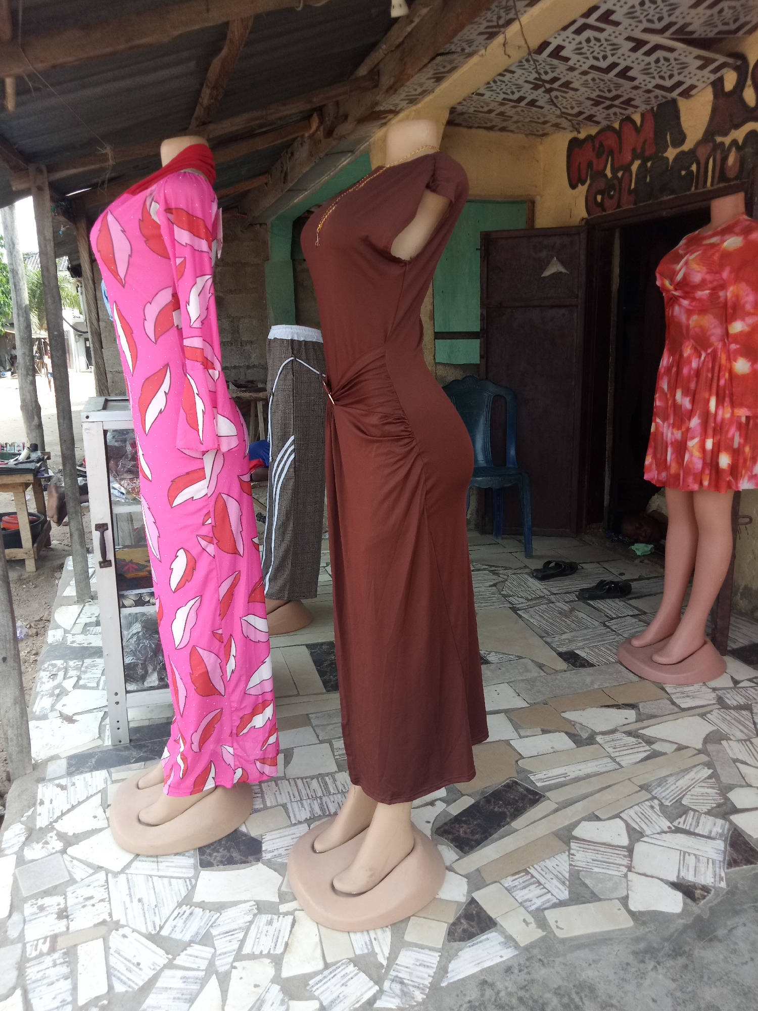 A medium shot captures a row of mannequins displaying clothing outside a shop. The mannequins are positioned on a patterned tiled floor with a rough, unfinished wooden overhang above. The mannequin on the left wears a long-sleeved pink dress with a repeating white and red leaf-like pattern. The middle mannequin is dressed in a long, brown, ruched dress. To the right, another mannequin displays a short, orange and pink floral print dress. Behind the mannequins, a doorway leads into a dimly lit interior. The shop signage above the doorway is partially obscured but reads "MAMA COLLECTION". The environment appears to be a street-side market or stall in an outdoor setting.
