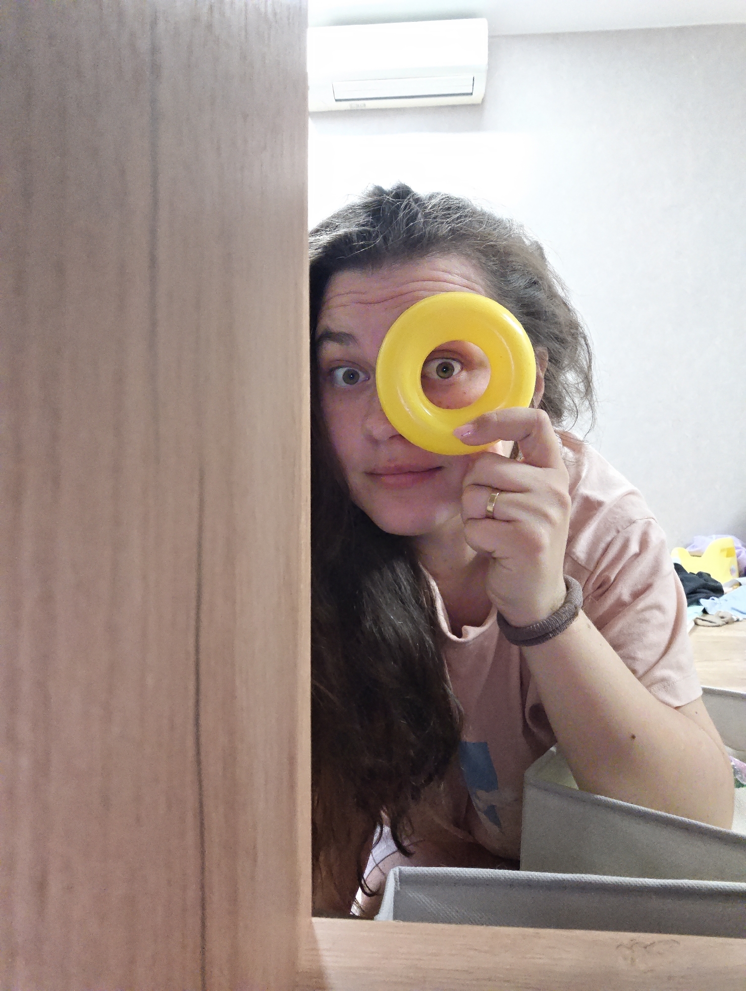 A woman is partially visible in a room, peeking from behind a light brown, wooden surface. She is holding a yellow, plastic ring up to her left eye, peering through the hole. Her dark, wavy hair is partially obscuring her face. She is wearing a light pink t-shirt and a dark elastic wristband. A gold ring is visible on her left ring finger.
In the background, there is a light-colored wall and an air conditioning unit mounted high on the wall. Clothes and other objects are scattered on the floor in the background.
The scene is located in Chaikovsky, Russia.