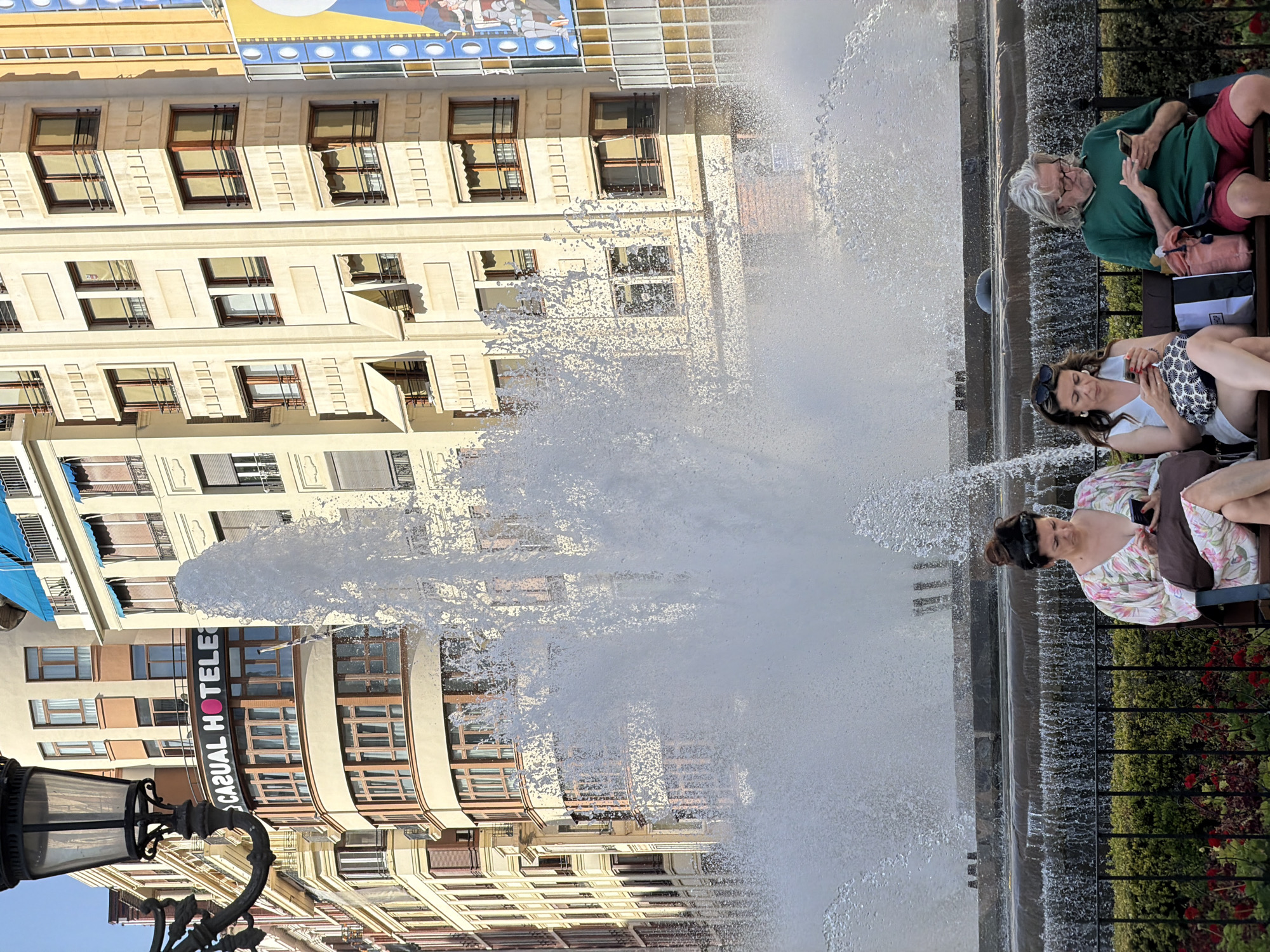 In València, Spain, a large, tiered fountain dominates the scene, with water spraying upwards in a dynamic display. Three individuals are seated on a dark bench in front of the fountain, each engrossed in their mobile phones. The person on the left wears a floral-patterned dress and has their hair pulled up. The person in the middle wears a white top and sunglasses perched on their head. The person on the right has long, gray hair and wears a green t-shirt and red shorts. Behind the fountain, a multi-story building with cream-colored facades and numerous windows rises. A sign on the building reads "CASUAL HOTELES" with a pink circular accent. The overall impression is a public urban space with a focus on leisure and technology.