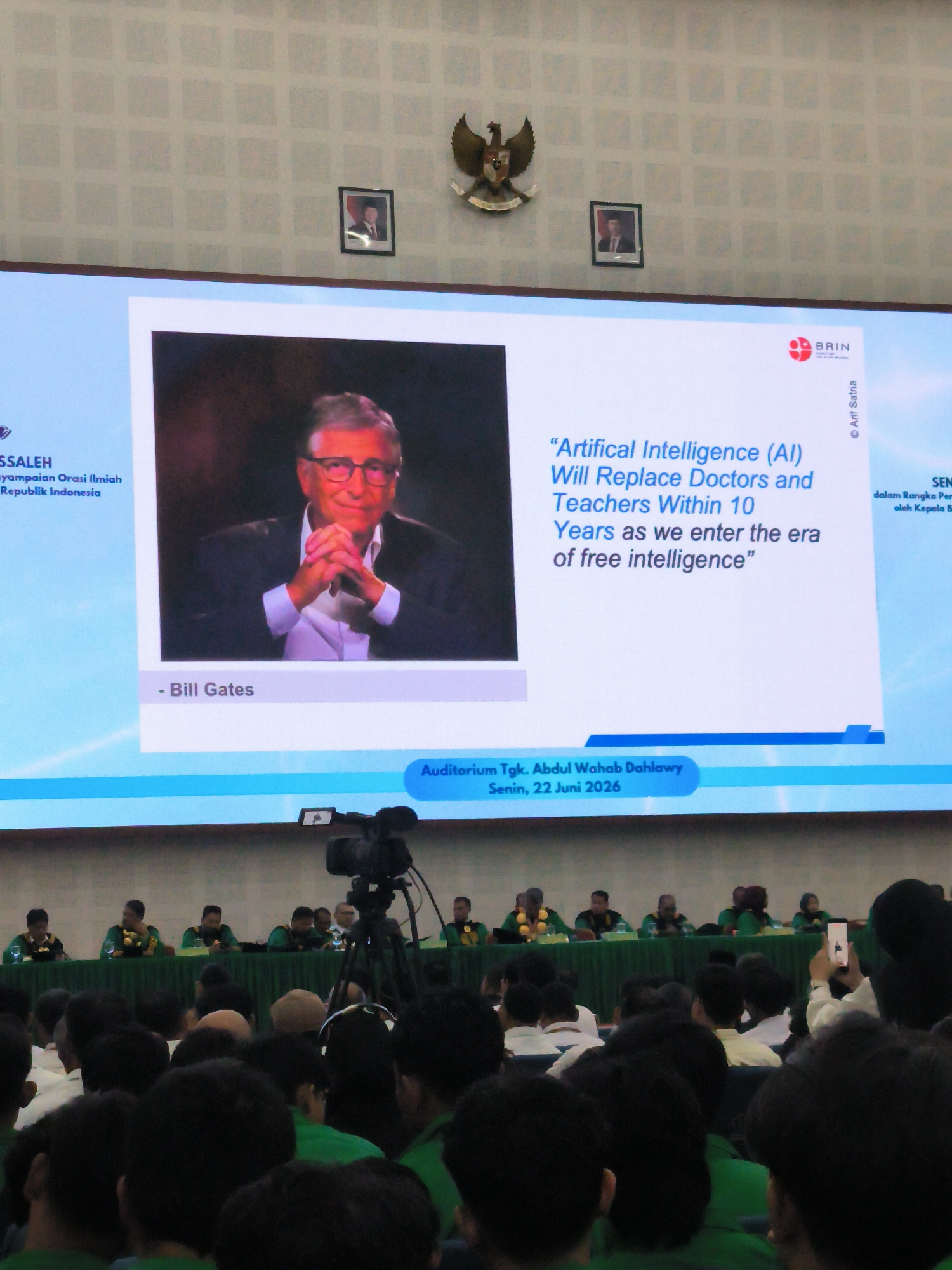 The image shows an indoor event in the Auditorium Tgk. Abdul Wahab Dalawiy in Reuleut Timu, Indonesia, on Monday, June 22, 2026. A large screen displays a presentation with a portrait of Bill Gates and a quote about artificial intelligence. The audience, seated in the foreground, is largely composed of young adults, many wearing green shirts. A video camera on a tripod is positioned to record the event. In the background, a stage is set up with several individuals seated behind a green tablecloth, facing the audience. The walls of the venue are decorated with patterned wallpaper, and two framed portraits and a national emblem are visible near the ceiling.