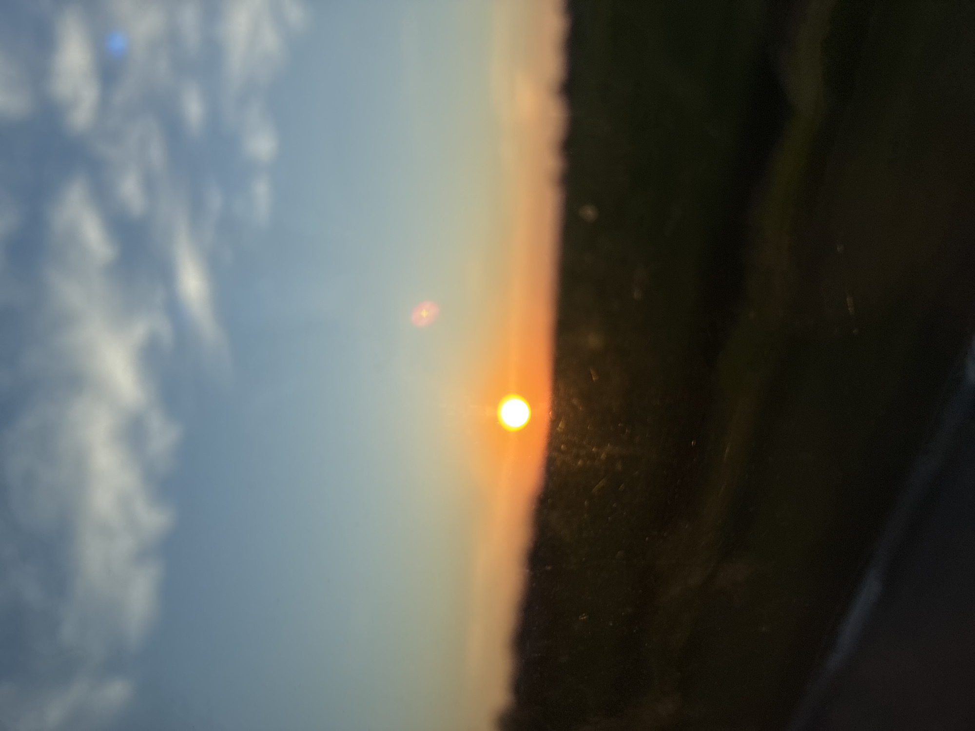 The image, when viewed rotated 90 degrees clockwise, presents a serene horizontal landscape scene during either sunset or sunrise. The sky dominates the upper and central portions, transitioning from a light blue with scattered white, fluffy clouds on the left to a warm, soft orange and pink gradient closer to the horizon on the right.

A prominent, bright yellow-orange sun, appearing as a distinct circular orb, is positioned slightly left of the image's vertical center, near the horizon. Several faint lens flares or reflections are visible: a small, reddish-orange glow to the left of the sun, and a subtle blue artifact within the clouds in the upper left.

Below the vibrant sky, a dark, silhouetted line stretches across the horizon, suggesting a distant tree line or hilly terrain. The right side and bottom portion of the image are occupied by a dark, slightly reflective surface, possibly a car window or similar glass, which frames the natural vista and adds depth to the foreground.

The scene conveys a peaceful atmosphere, characteristic of the transition periods of the day. The absence of specific landmarks, beyond the general outline of trees or hills, suggests a natural, open environment. The location context indicates this tranquil scene is set in Harlets, Bulgaria. There is no visible text or direct human activity depicted.