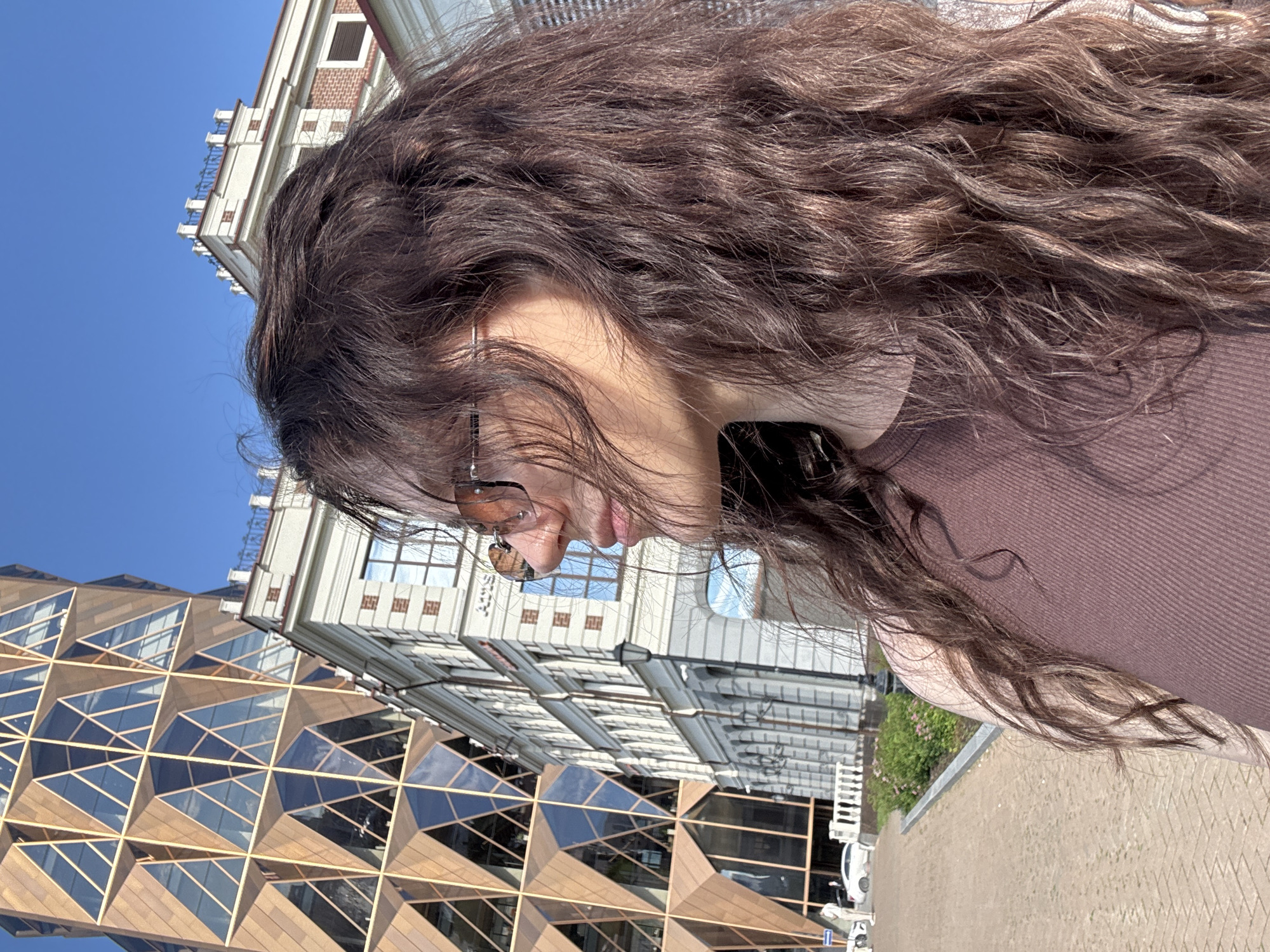 A young woman with long, wavy brown hair is captured in a profile view, looking down. She is wearing sunglasses with thin, tinted lenses and a mauve, ribbed top. Her hair is partially obscuring her face and shoulders, with some strands falling across her cheek and in front of the sunglasses.
The background features a clear blue sky and a modern building with a distinctive, geometric facade composed of glass and bronze-colored frames. Adjacent to this is a taller, classical-style building with white and red brick detailing, arched windows, and decorative white railings along its upper levels. The scene appears to be an urban environment, possibly a city street or plaza, given the presence of buildings and paving stones in the foreground.
The lighting suggests it is daytime, likely mid-morning or afternoon, with bright sunlight casting shadows and highlighting the textures of the buildings and the woman's hair. The overall impression is one of a casual outdoor moment, possibly in Yekaterinburg, Russia, given the location context. No discernible text is visible in the image.