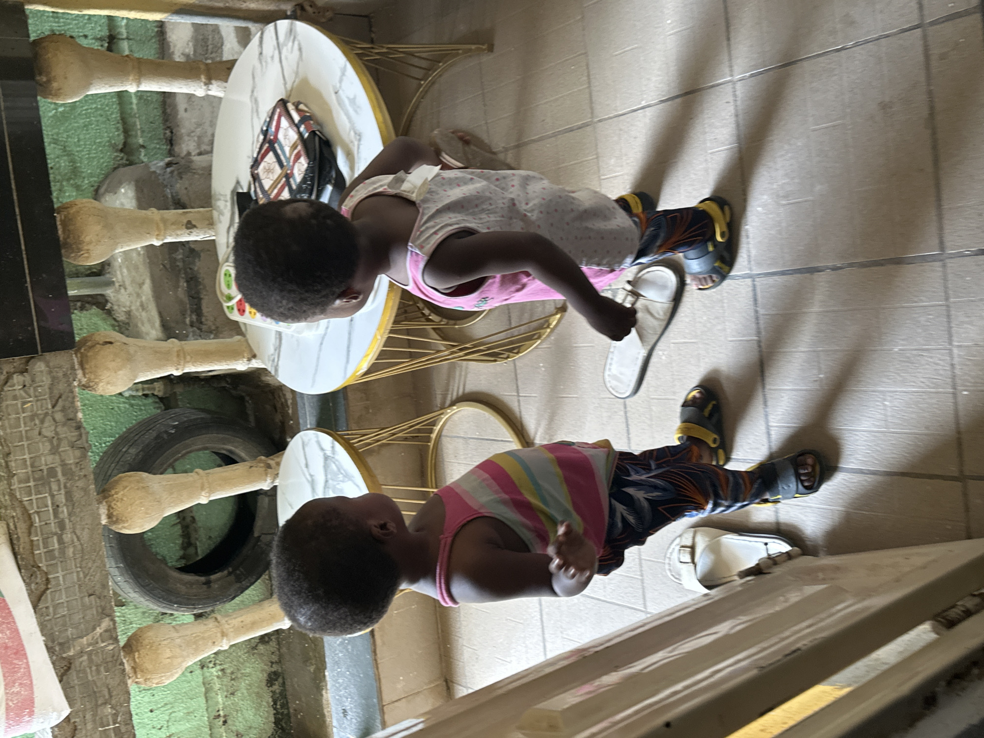 Two young children, their backs to the camera, stand on a tiled floor in what appears to be an outdoor or semi-outdoor space. The child on the left wears a striped tank top and patterned pants. The child on the right wears a white top with pink polka dots over a pink undergarment and patterned pants, and appears to be wearing yellow and black sandals. White flip-flops are visible on the floor near both children. In the background, a round table with a marble-patterned top is visible, holding a colorful object and a patterned clutch or wallet. Another round table is partially visible to the left. Behind the tables, a green wall is visible, with several beige, decorative posts extending from it. A black tire is leaning against the wall. This scene is located in Narayi, Nigeria.