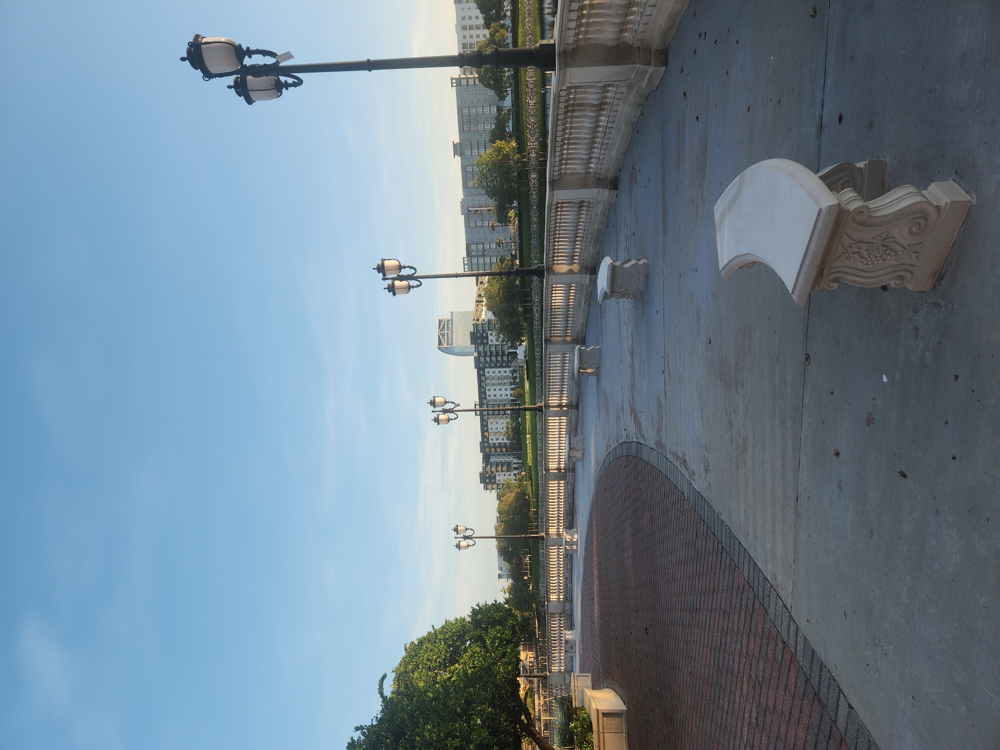 The image shows a waterfront promenade in Altamonte Springs, Florida, USA. A paved walkway curves along a body of water, bordered by a decorative stone balustrade. Several ornate street lamps with multiple lights are spaced along the promenade, and there are several stone benches with decorative bases. To the left, a section of the walkway is paved with red bricks, and a large green tree with dense foliage is visible. In the background, across the water, are several multi-story buildings, including a modern high-rise with a curved facade. The sky is clear and blue, indicating it is daytime.