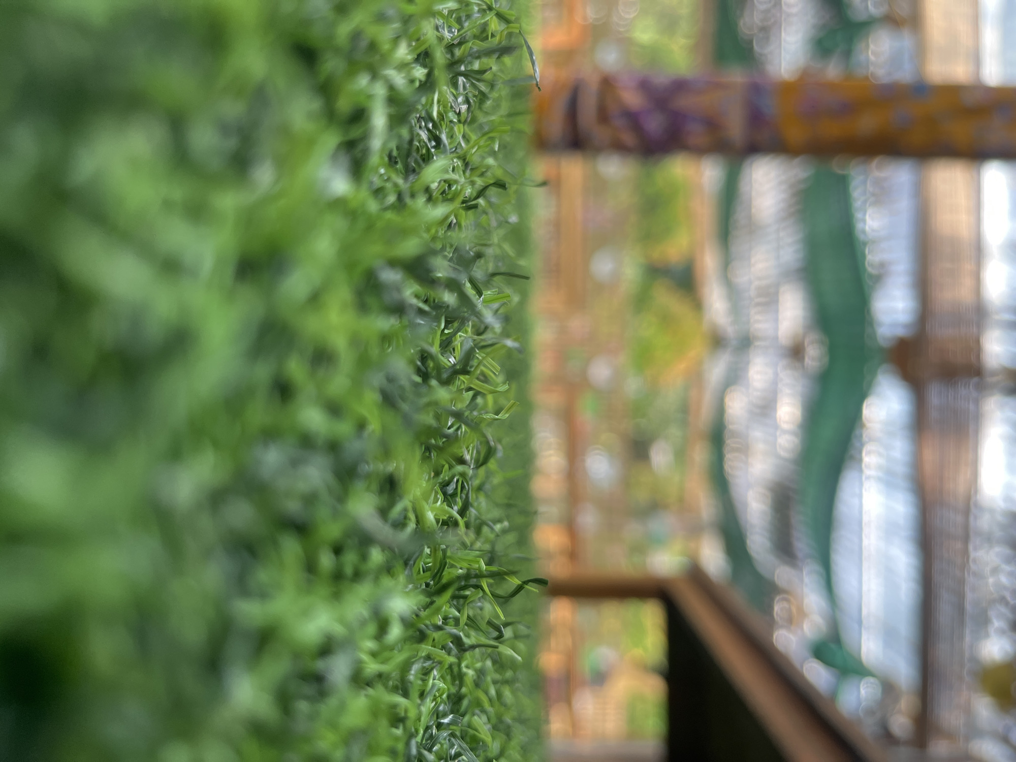 A low-angle shot showcases a patch of artificial green grass in what appears to be an outdoor seating area. The grass blades are detailed and in focus in the foreground, with a single blade slightly curved towards the left. In the background, the scene is blurred. To the left, a tall, decorative post with yellow and purple patterns is visible. Further back, a wooden structure, possibly a roof or pergola, is suggested, with green fabric draped beneath it. The overall environment suggests a public space or an outdoor cafe in Tangerang, Indonesia, likely designed for leisure or dining. No people are present in the image.