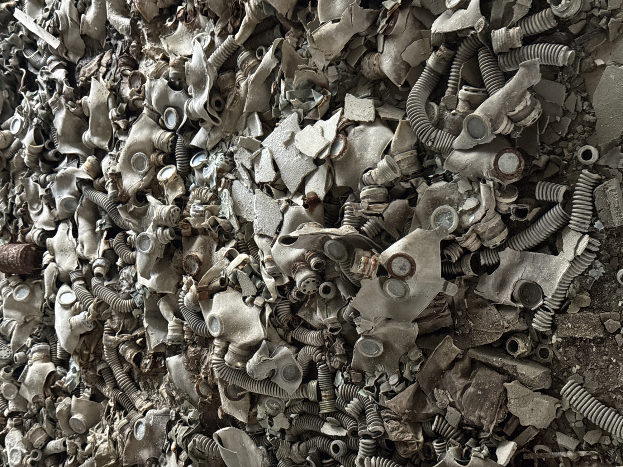 The image depicts a dense pile of discarded and broken gas masks in what appears to be an interior space. The masks are predominantly gray and appear aged or damaged, with visible signs of wear, cracks, and deterioration. Some masks are partially disassembled. Corrugated hoses and canister components are interspersed with the mask faces. The background and surrounding environment are obscured by the density of the pile, and the overall setting suggests a desolate or abandoned location. The location is possibly in the city of Prypiat, Ukraine.