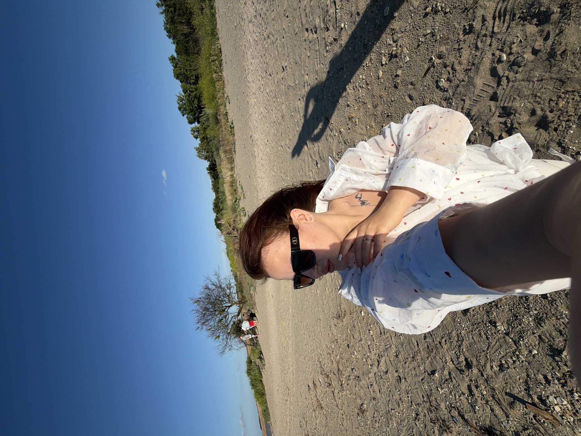 The image depicts a young woman with brown hair, wearing a white shirt with colorful heart patterns and black sunglasses, posing for a selfie on a sandy beach. She is sitting with her legs crossed, and her left arm is extended towards the camera. A tattoo is visible on her collarbone. In the background, a bare tree stands on a sandy dune, with a few people standing near its base. The scene is set under a clear blue sky, suggesting it is a sunny day. The location is identified as the city of Port-Katon, Russia. The sand is a mix of fine grains and small pebbles. The overall impression is a casual, outdoor moment.