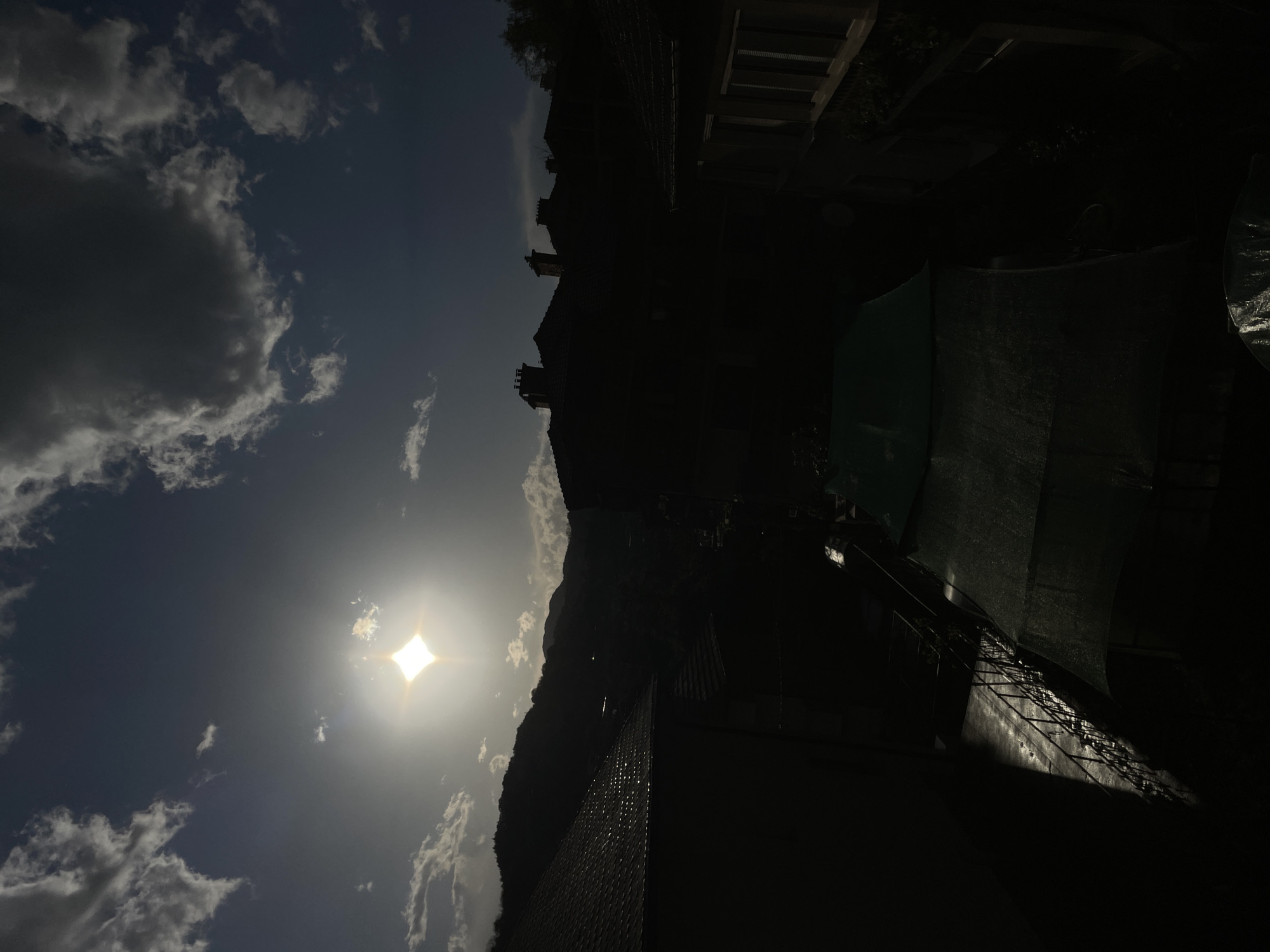 The image captures a silhouette of buildings and structures against a bright sky with clouds. The sun is prominently visible. The structures are not clearly discernible. The location is likely near or in a neighborhood based on the presence of buildings. The precise location within Zlatograd, Bulgaria, is impossible to determine.