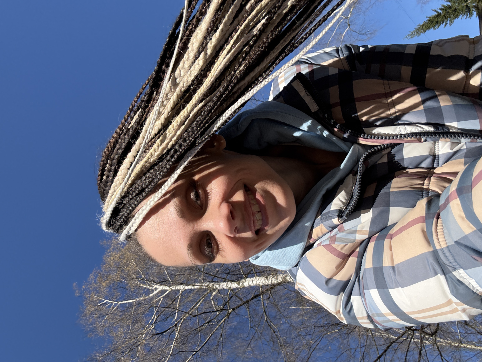 The image captures a selfie of a smiling person outdoors on a bright, sunny day. The subject has an elaborate hairstyle featuring numerous dark brown and light-colored (blonde/beige) braids, which extend upwards and are visible around their head. They are wearing a light blue hooded top under a plaid puffer jacket. The jacket has a distinct checkered pattern with colors including dark blue/grey, beige, brown, and light blue/white.
The person looks directly at the camera with a wide, cheerful smile, revealing their teeth. Strong sunlight casts shadows from their braids across their forehead and face, indicating bright conditions. The background consists predominantly of a clear, vibrant blue sky. Visible against the sky are the bare branches of deciduous trees, suggesting the season is late winter or early spring. In the upper right corner, a hint of evergreen foliage is also present.
The scene conveys a sense of happiness and enjoyment of the outdoors, likely in a park or natural setting. There is no visible text in the image. The location context is Rusanovo, Russia.