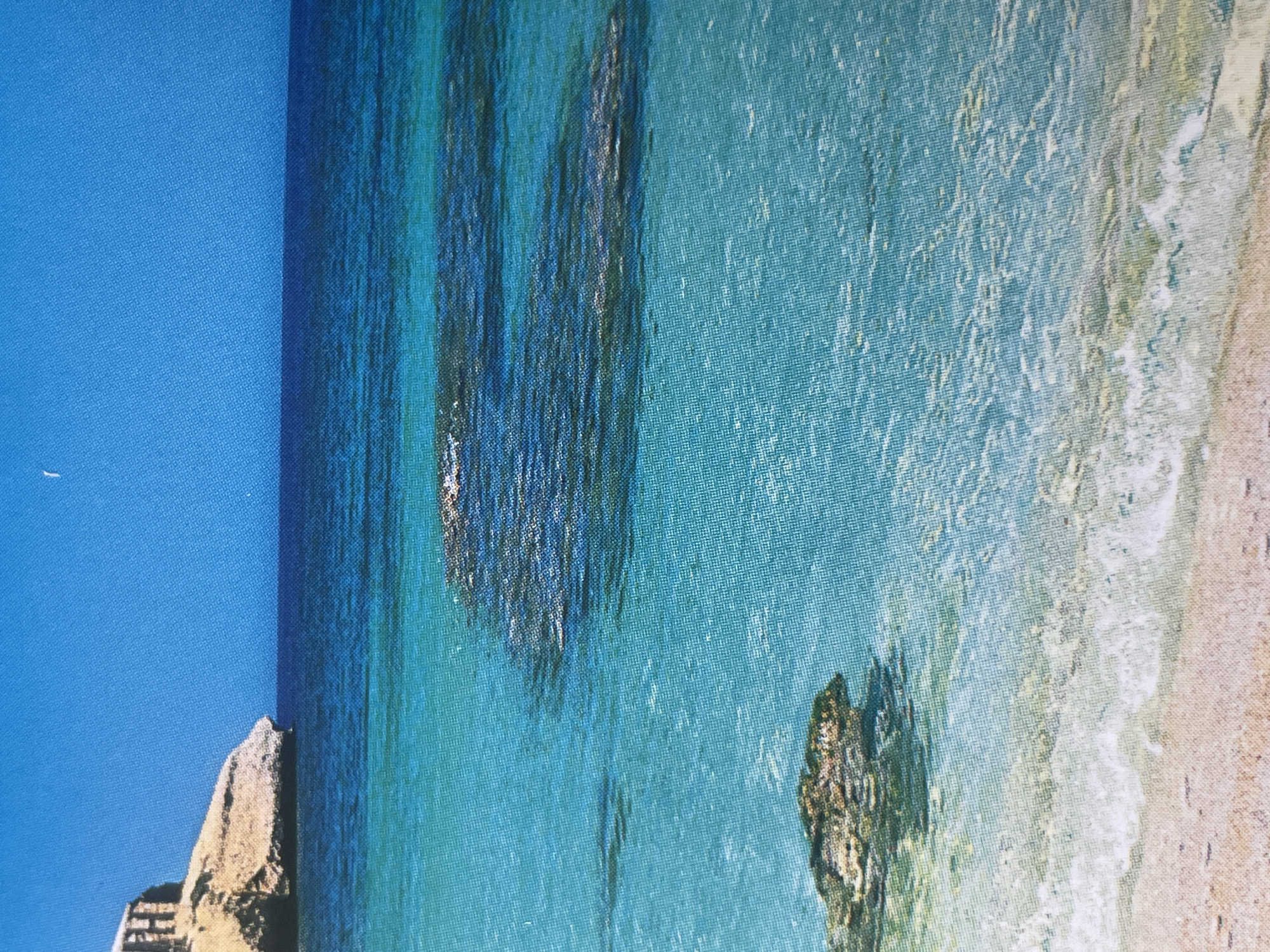This is a photograph of a coastal landscape featuring clear turquoise water lapping onto a sandy beach. In the background, a rocky cliff with a structure on top juts out into the sea under a bright blue sky. There are several submerged rocks visible in the water. The image does not depict any elements related to the city of Sofia, Bulgaria.