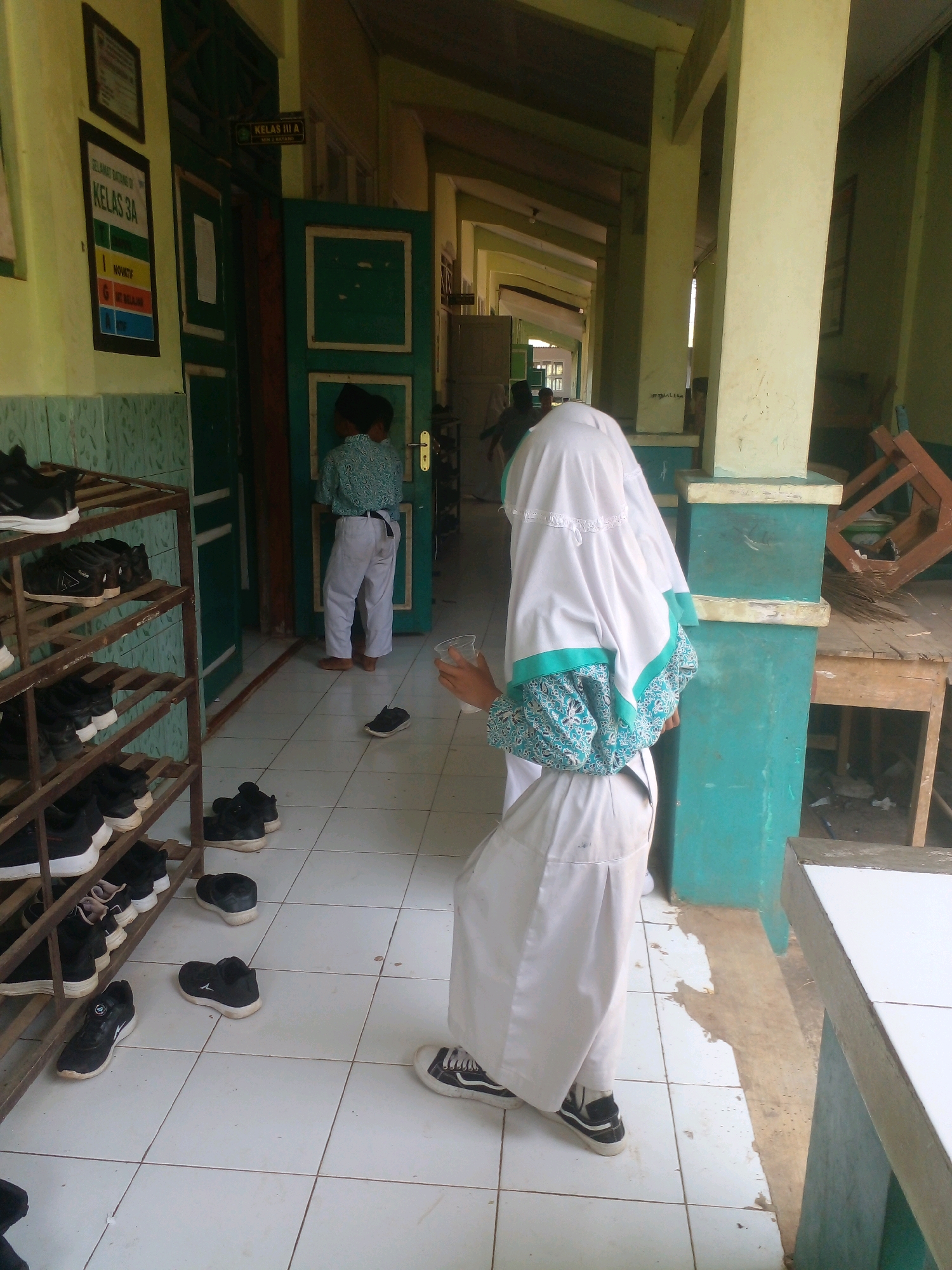The image depicts an indoor scene, likely a school hallway in Banyuputih, Indonesia. A young person, wearing a white hijab and a teal-patterned garment, is in the foreground, facing away from the camera and holding a clear plastic cup. Their attire suggests they are likely a student. In the middle ground, another student, wearing a teal patterned shirt and white pants, stands near a green door, looking into the classroom. The hallway features tiled flooring and a shoe rack on the left filled with black shoes. Signs are visible on the wall, one reading "KELAS 3 A" (Class 3A). The environment appears to be a school building, with green doors, light-colored walls, and pillars. To the right, a wooden table and chair are visible, suggesting a common area or classroom space.
