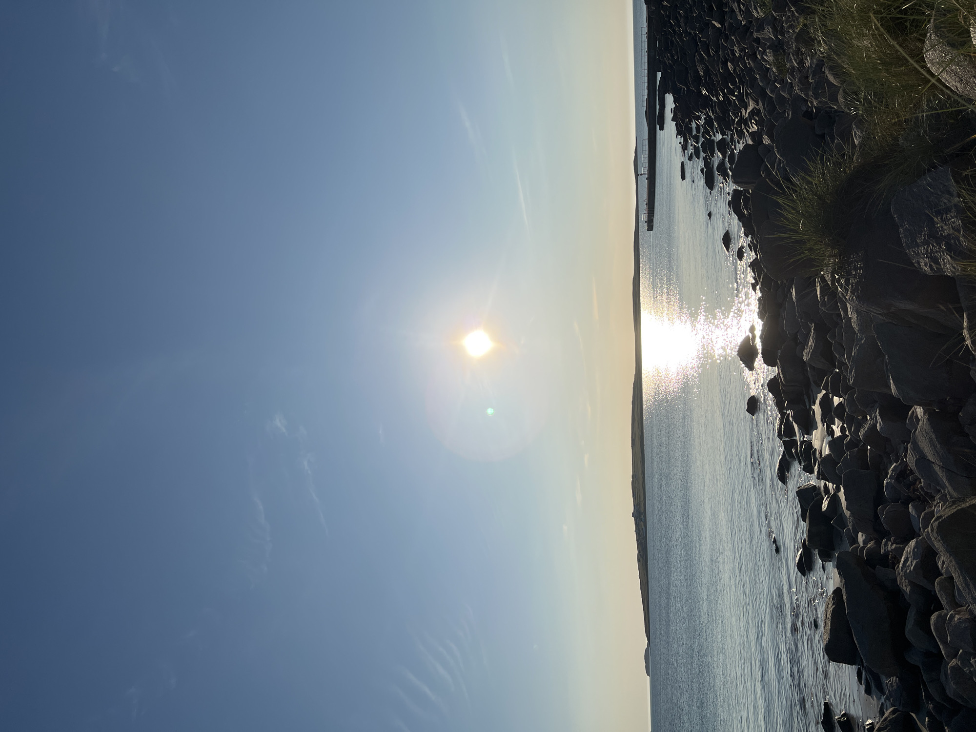 The image depicts a sunlit seascape on a clear day, likely in Lunniaghbeg, Ireland. The foreground is a rocky shoreline with scattered grasses. The sea is calm, reflecting the bright sun which is visible in the upper left quadrant of the image, creating a glinting path across the water. In the distance, a long, thin island with some structures on it is visible on the left horizon. A pier extends into the water on the right side of the frame, with a single, small figure standing on it. The sky is a clear, pale blue with faint wispy clouds.
