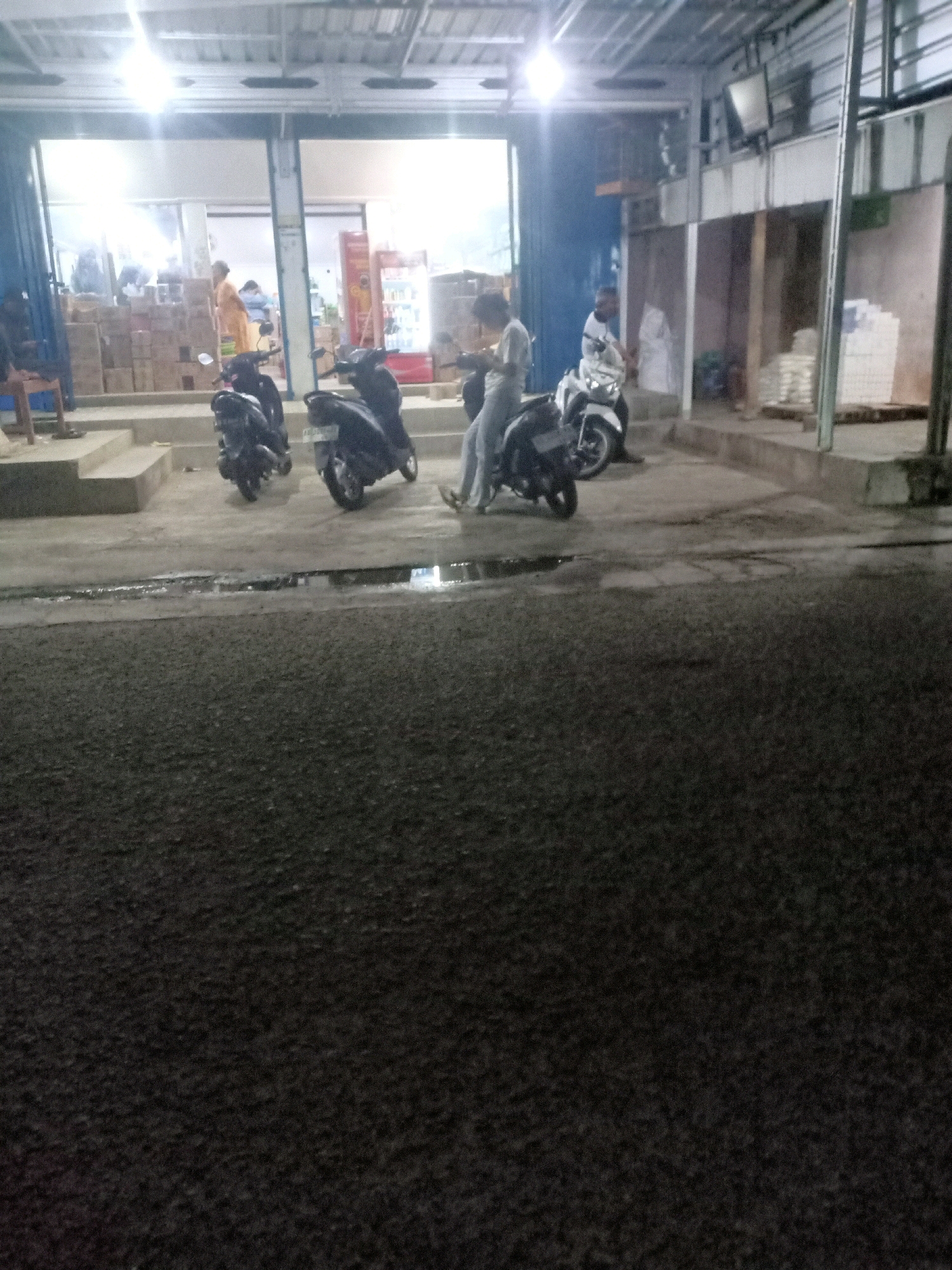 A low-angle, slightly blurry, medium shot captured at night shows a street scene in front of a shop in Tremes, Indonesia. The shop's interior is illuminated, revealing stacks of boxes and several people. Outside the shop, three parked motorbikes are visible. Two are dark-colored and in the foreground. A lighter colored motorbike with a rider is positioned further back. The foreground of the image is dominated by a dark, wet asphalt surface, suggesting recent rain. The overall impression is of a busy commercial area at night.