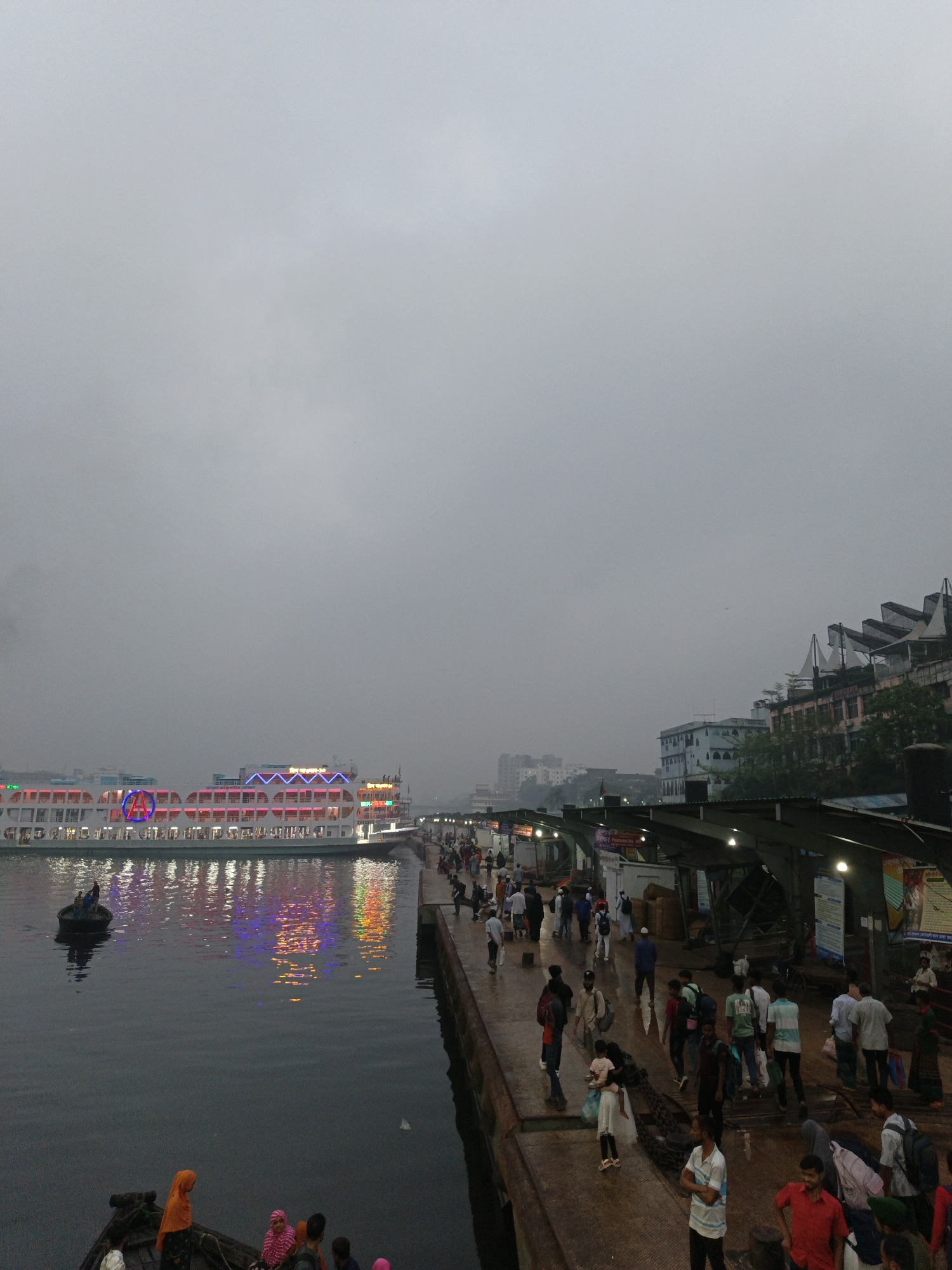The image depicts a riverside scene in Dhaka, Bangladesh, likely near Sadarghat, a major river port on the Buriganga River. It's overcast, suggesting either early morning or a cloudy day. A large passenger ferry is docked parallel to a concrete pier, its outline illuminated with colorful lights that reflect in the water. Several smaller boats are visible in the foreground.
A large number of people are on the pier, including individuals and families. They are dressed in a variety of clothing styles. Some are walking, some are standing, and some appear to be interacting. Buildings and structures line the opposite side of the pier, including covered stalls or shops, and taller structures beyond. The scene suggests a bustling river port environment.