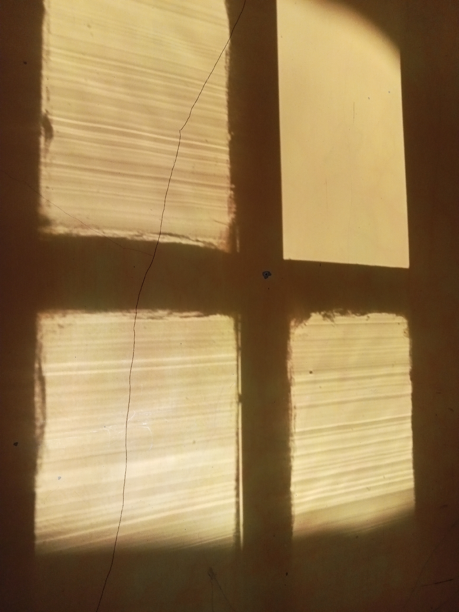 This is a close-up, low-angle shot of light patterns on a wall. The light is cast by a window with horizontal blinds, creating distinct rectangular shapes and stripes on the wall. The colors are predominantly warm tones of yellow and brown, with areas of shadow. A thin, dark crack runs vertically through the center of the image, diagonally across one of the illuminated rectangular sections. There are a few small, dark specks on the wall. The scene appears to be indoors, possibly in a room in Pancurwening, Indonesia, where sunlight is entering through a window and casting shadows. There are no people or discernible objects other than the wall, the light patterns, and the crack.