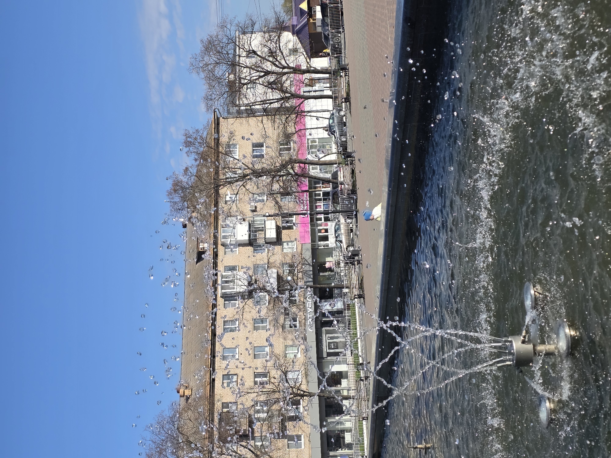 The image shows a view from a fountain in the city of Chaikovsky, Russia. The fountain features a multi-nozzle spray system. Behind the fountain is a four-story brick building, with several trees in front of it. A sidewalk runs in front of the building, and behind that, another building is partially visible. The sky is blue with some clouds.