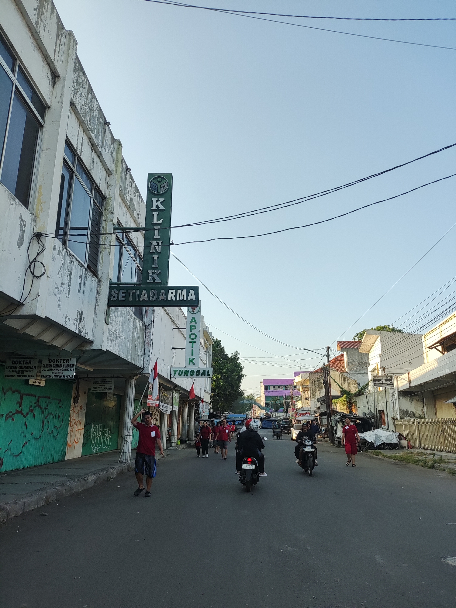 The image captures a street scene in Cirebon, Indonesia, under a clear, bright sky, suggesting it's early morning or late afternoon. A building with faded white paint and large windows is on the left, with signs indicating "KLINIK SETIADARMA" and "APOTIK TUNGGAL." The clinic sign is green with the word "KLINIK" stacked vertically, and "SETIADARMA" horizontally below it. A group of people, some wearing red shirts, are walking on the sidewalk and in the street. Several motorcycles are present, with some people riding them. The street is paved and appears to be a local thoroughfare with various shops and buildings lining both sides. Trees are visible in the background, and electrical wires crisscross overhead. One man in the foreground is walking on the road, raising his right hand. Another man in a red shirt is walking on the sidewalk. The overall impression is of a typical urban street in Indonesia.
