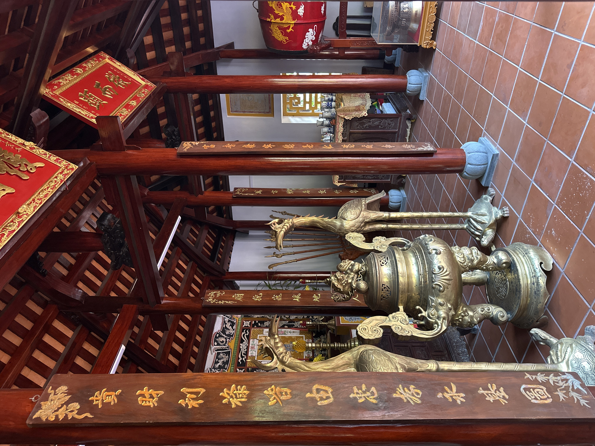 The image depicts the interior of a temple, likely a traditional Vietnamese one, with a focus on its architectural elements and decorative objects. The structure features dark red wooden pillars supporting a beamed ceiling with a visible wooden roof structure. The floor is tiled with reddish-brown square tiles.
Prominently featured are several large, ornate brass objects. To the left, a tall, slender brass statue of a bird-like creature with reptilian feet stands beside a wooden post inscribed with vertical golden Chinese characters. In the center, a large, elaborate brass incense burner rests on a base adorned with dragon motifs and sits on the tiled floor. Another brass bird statue, similar to the first, is positioned to the right of the incense burner. Further back, in the corner of the room, a large red drum with a golden dragon design is visible.
The background reveals more temple decor, including framed artwork or scrolls on the walls, a table with a patterned cloth laden with decorative vases, and what appear to be weapons or staffs arranged upright. The overall atmosphere suggests a place of worship or historical significance, with rich cultural and religious symbolism. The presence of Chinese characters on the wooden posts indicates a historical connection to Chinese culture, which is prevalent in Vietnamese temples. This image was taken in the city of Đà Nẵng, Vietnam.