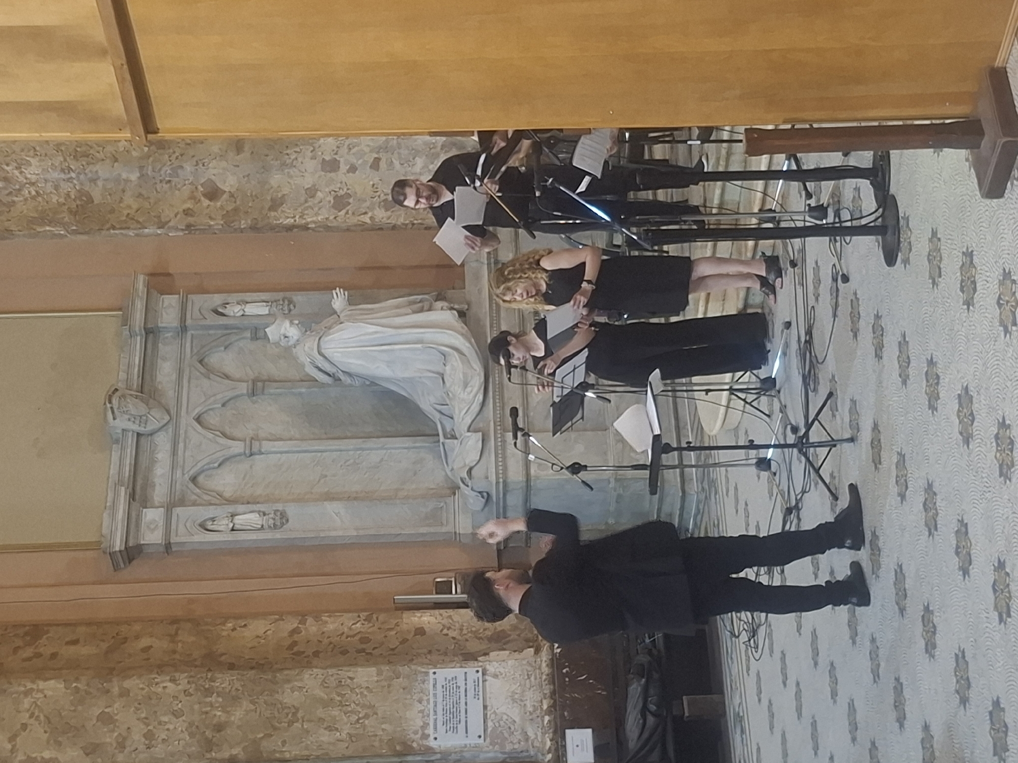 Inside a church, possibly the Catedral Metropolitana in Buenos Aires, Argentina, there is a musical performance. A conductor in black stands facing three musicians. The musicians, two women and a man, are playing stringed instruments. They are standing with music stands, and they are reading sheet music. The church has a patterned floor, tall walls, and a statue.