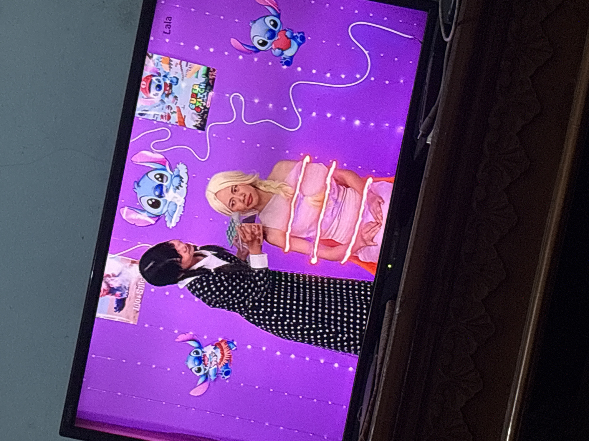 A television screen displays a purple background with scattered white dots, resembling stars. Two young women are visible on the screen. One, with dark hair and wearing a black and white polka dot dress and white collar, stands on the left, interacting with the other. The second woman, with blonde hair in a braid and wearing a pink strapless dress wrapped with white string, is seated on the right. Cartoon characters of Stitch, a blue alien from Disney's Lilo & Stitch, are placed around the screen. One Stitch is on the upper left, another is on the lower right holding a red heart, and a third is on the upper right wearing a red hat and blue overalls, appearing to be from a "Super Stitch" game. A small image with the text "Woah Stitch" is also visible on the upper left. The text "Lala" appears on the upper right. The scene is captured from an angle that shows the edge of the television and the wooden stand it rests on. The overall lighting suggests an indoor setting, likely a living room, with the image focused on the television screen. There is no discernible indication of the location being Palimanan, Indonesia.